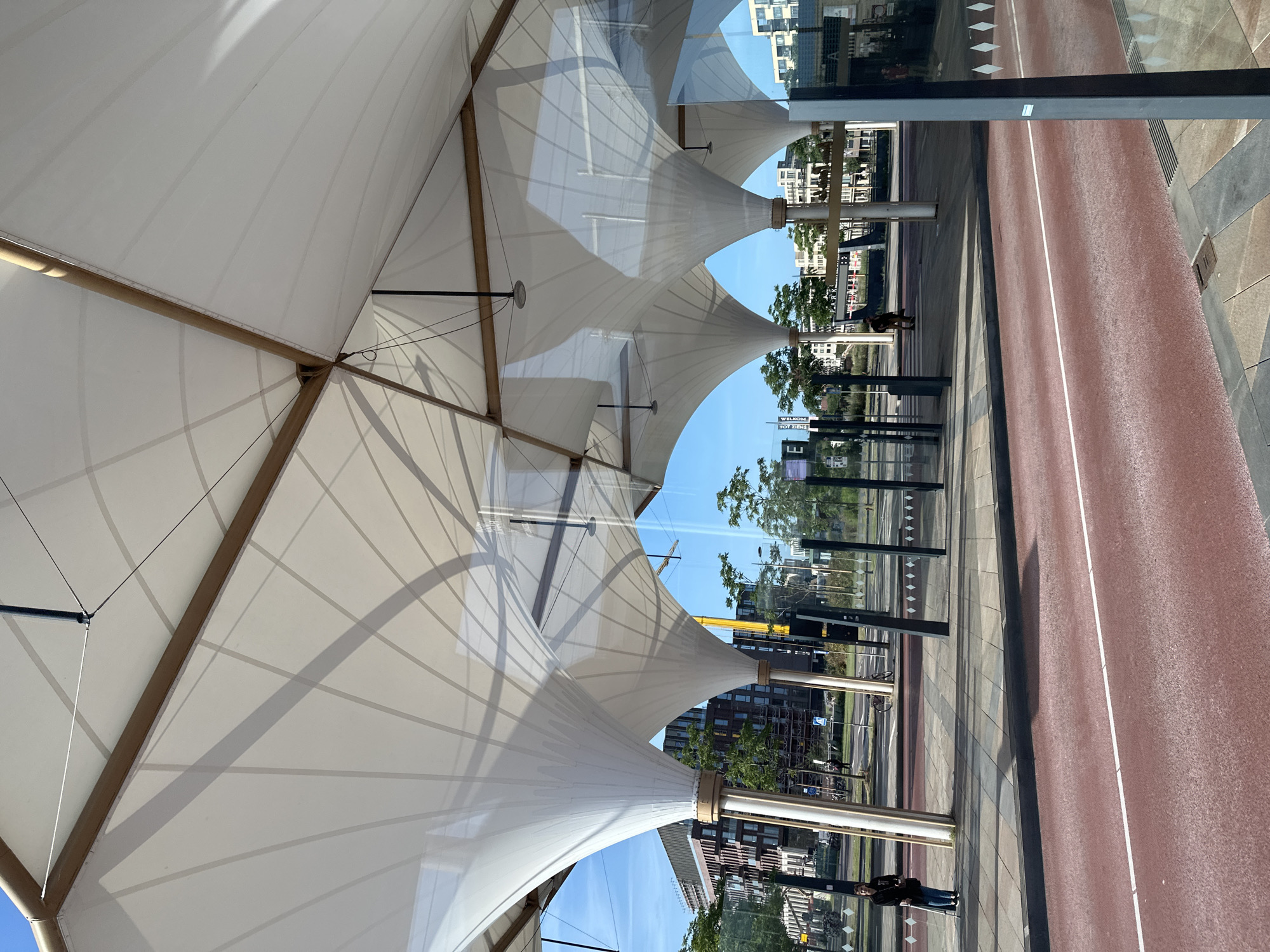 The image depicts a modern bus stop shelter with a large, white, tensile fabric roof supported by wooden beams and white pillars. The shelter is situated on a street in Utrecht, Netherlands. The roof has a geometric design with a series of curved segments that create a visually striking pattern. Sunlight filters through the translucent fabric, casting shadows on the ground below.
In the foreground, a red-colored road surface with a white line runs horizontally across the frame, indicating a traffic lane. To the left of the road, a grey sidewalk is visible, with diamond-shaped patterns etched into its surface. Several dark, slender poles stand along the sidewalk, likely part of the bus stop infrastructure.
A woman, dressed in dark clothing and carrying a bag, stands on the sidewalk near the left edge of the image, leaning on a walking stick. Further down the sidewalk, other individuals are visible, some standing and others walking. In the background, modern buildings with numerous windows and balconies line the street, interspersed with trees. A yellow construction crane is also visible in the middle ground. The sky is clear and blue. The scene appears to be a bright, sunny day.