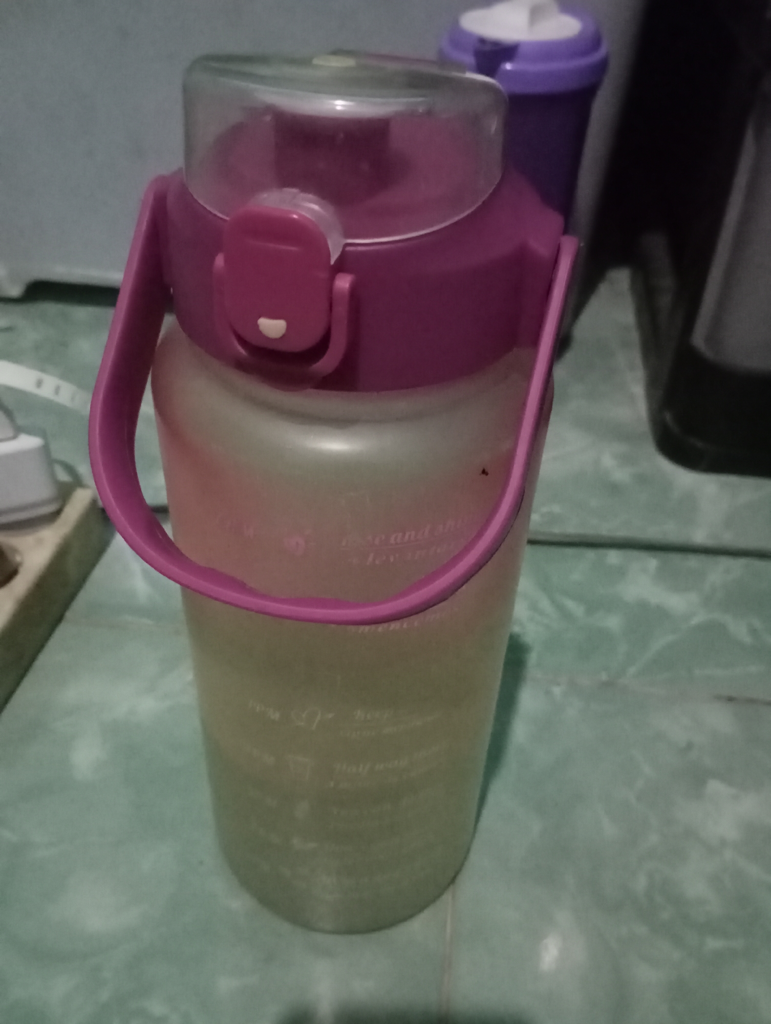 A purple-lidded water bottle with a clear plastic lid sits on a green tiled surface. The bottle is translucent and appears to contain a light-colored liquid. The bottle has a pink handle attached to the lid. In the background, a purple and white container is visible. To the left of the water bottle, a white electrical outlet is partially visible on a light-colored surface. The lighting is dim, creating shadows. The scene appears to be indoors.
