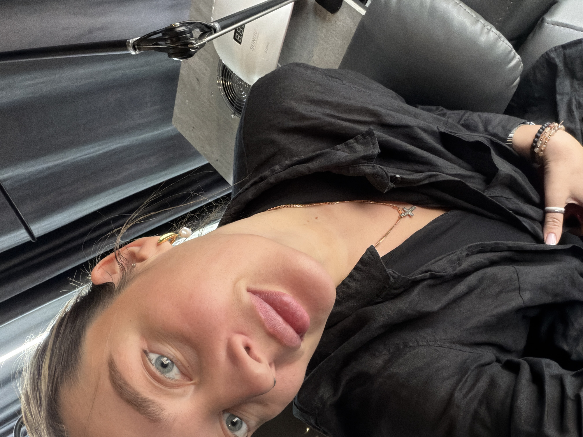 The image shows a young woman's face, tilted towards the camera, with blue eyes and fair skin. She is wearing a black shirt, and a gold necklace is visible around her neck. She has multiple bracelets on her wrist. A nail lamp is visible near her. The background suggests an interior setting. There is no indication of the city or location in the city of Moscow, Russia.
