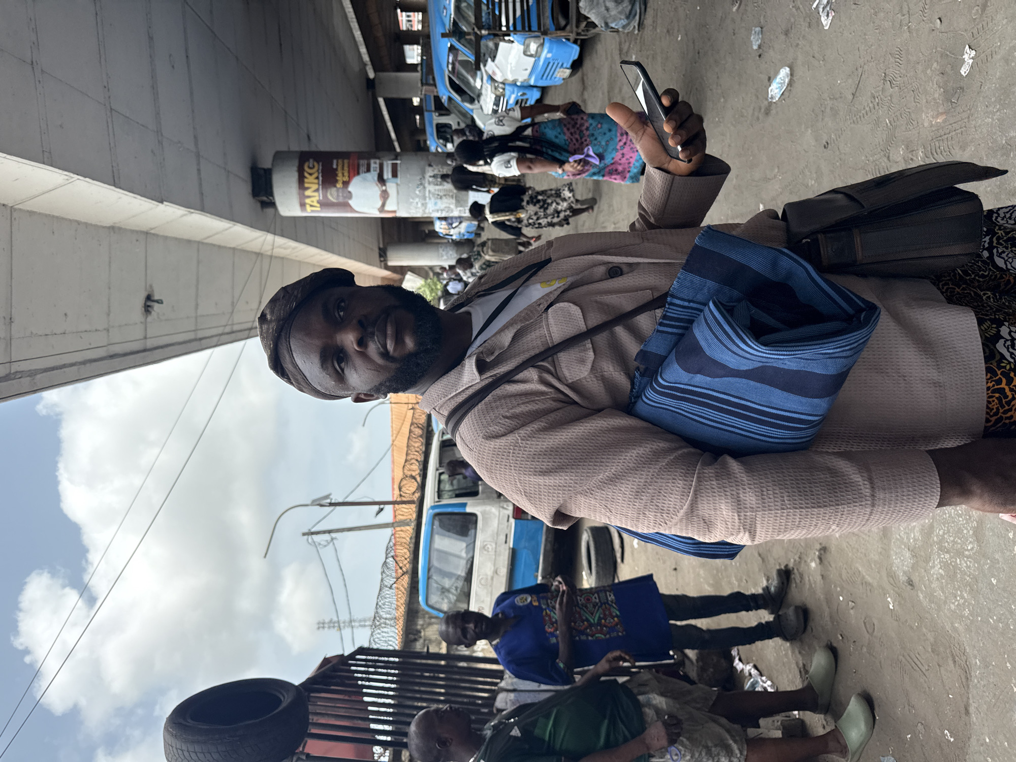 A man holds a smartphone in a crowded outdoor setting likely in Lagos, Nigeria. The man is middle-aged with a beard, wearing a cap and a light brown textured jacket over a patterned shirt. He has two bags slung over his shoulders: one dark leather and one blue striped cloth.
In the background, other people walk and sit along a concrete ground. There are blue and white public transportation vehicles. A large advertisement is affixed to a column above the scene, and a tire sits atop a metal structure. A chain-link fence is visible behind some people. The sky is cloudy.
