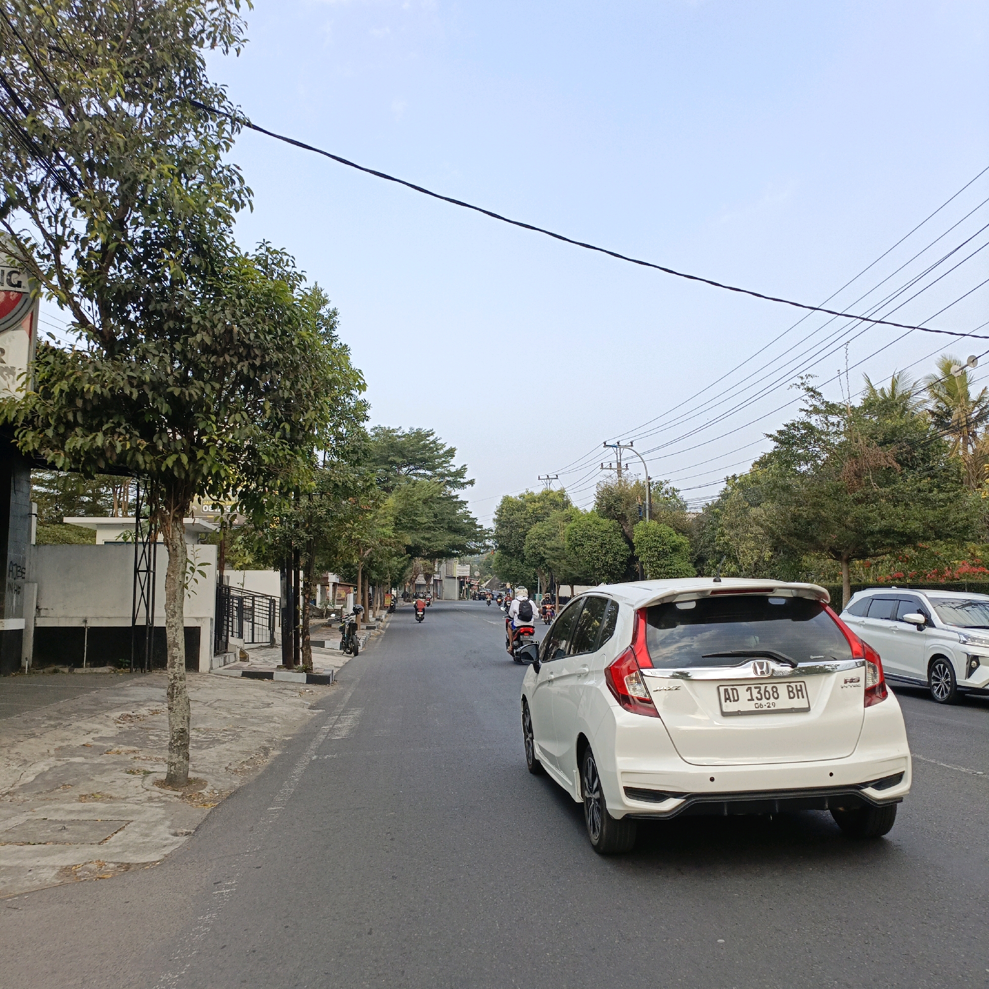 The image depicts a street scene in Salatiga, Indonesia. In the foreground, a white Honda Jazz is parked on the right side of the road, facing away from the camera. To the left, a tree-lined sidewalk runs alongside a building with a grey facade. The street is paved with asphalt and shows faint white lane markings. Several motorcycles are visible on the road, some carrying passengers, indicating moderate traffic. The background is filled with lush green trees and overhead power lines, characteristic of a tropical urban environment. The sky is clear and pale blue, suggesting it is daytime.