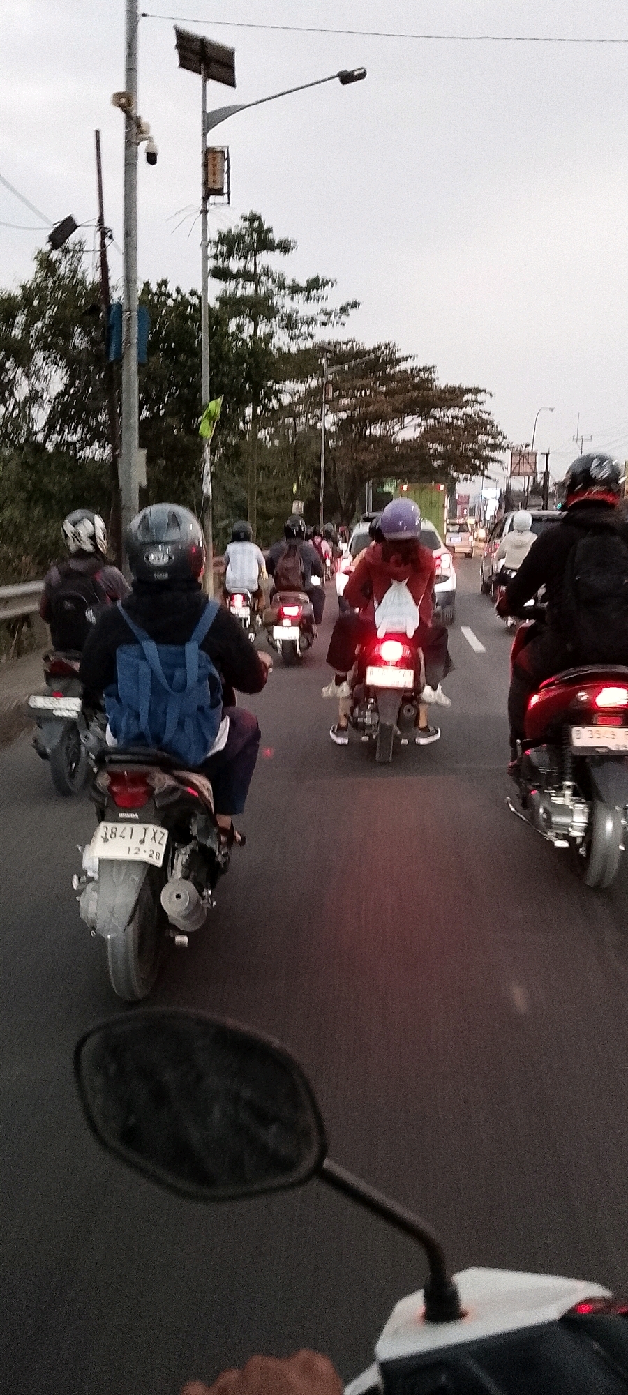 The image depicts a traffic scene, likely on a road in Depok, Indonesia, during what appears to be late afternoon or early evening, given the overcast sky and the dim lighting. The perspective is from the rear of a scooter, looking forward at a line of motorcycles and a few cars.
In the foreground, a person wearing a black helmet with a black top and a blue backpack is riding a scooter. The scooter's license plate is partially visible, reading "3841 JXZ". To the left, another scooter is partially visible with a rider wearing a helmet with a checkered pattern.
Further ahead, several other scooters are visible. One scooter has a rider wearing a purple helmet and a maroon jacket. Another scooter to the right has a rider wearing a black helmet and a black jacket. Several other individuals on scooters and in cars are also seen in the middle ground, forming a traffic jam.
The environment includes trees lining the sides of the road and some overhead wires and streetlights, including a streetlight with a solar panel. The road surface is asphalt. The general atmosphere suggests a common urban commuting scenario with moderate traffic.