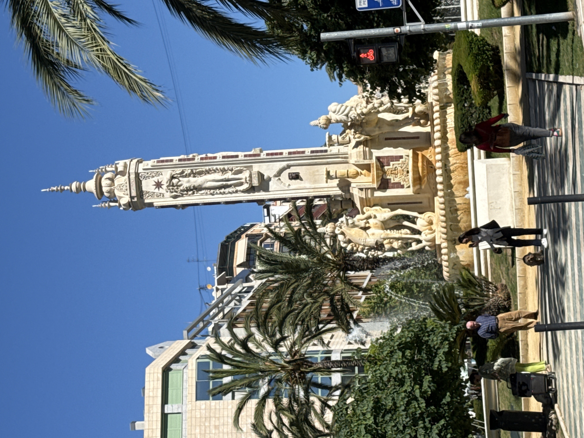 The image shows a view of the Fuente de la Aguadora, a fountain located in Valencia, Spain. The fountain is white and features sculptures of human figures and horses. There are palm trees and other greenery surrounding the fountain. People are walking on the sidewalk next to the fountain. The sky is blue. Buildings are visible in the background. A traffic light can be seen near the fountain.
