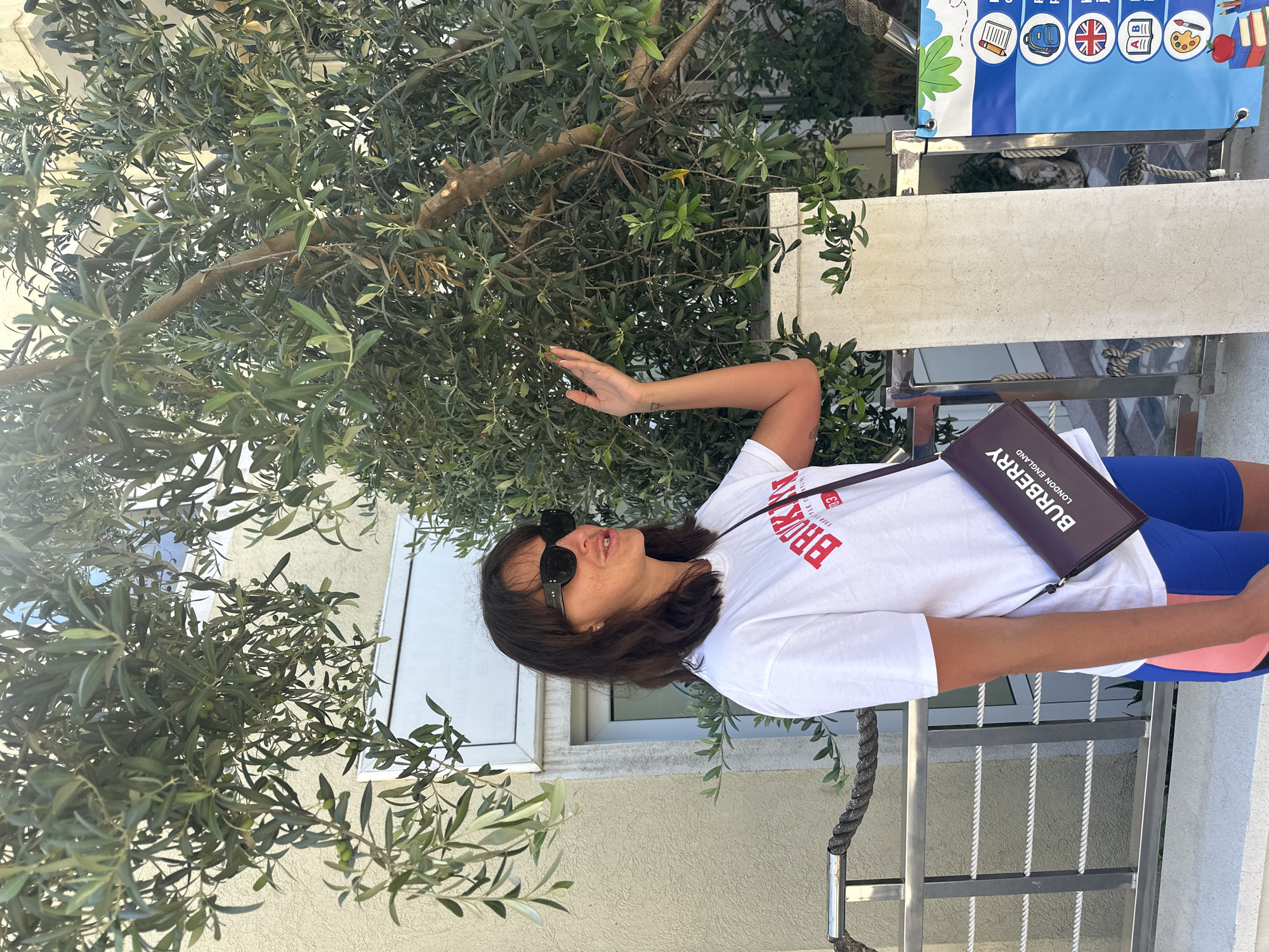 A woman is standing outdoors in front of an olive tree. She is wearing a white t-shirt with red lettering that says "BROOKLYN", sunglasses, and blue and pink shorts. A dark maroon purse with "BURBERRY LONDON ENGLAND" written on it is slung across her body. Her right hand is raised towards the olive tree. The background includes a building with light yellow siding, a metal railing with a rope, and a colorful sign with various icons. The scene appears to be in Budva, Montenegro, possibly near the Old Town or a coastal area.