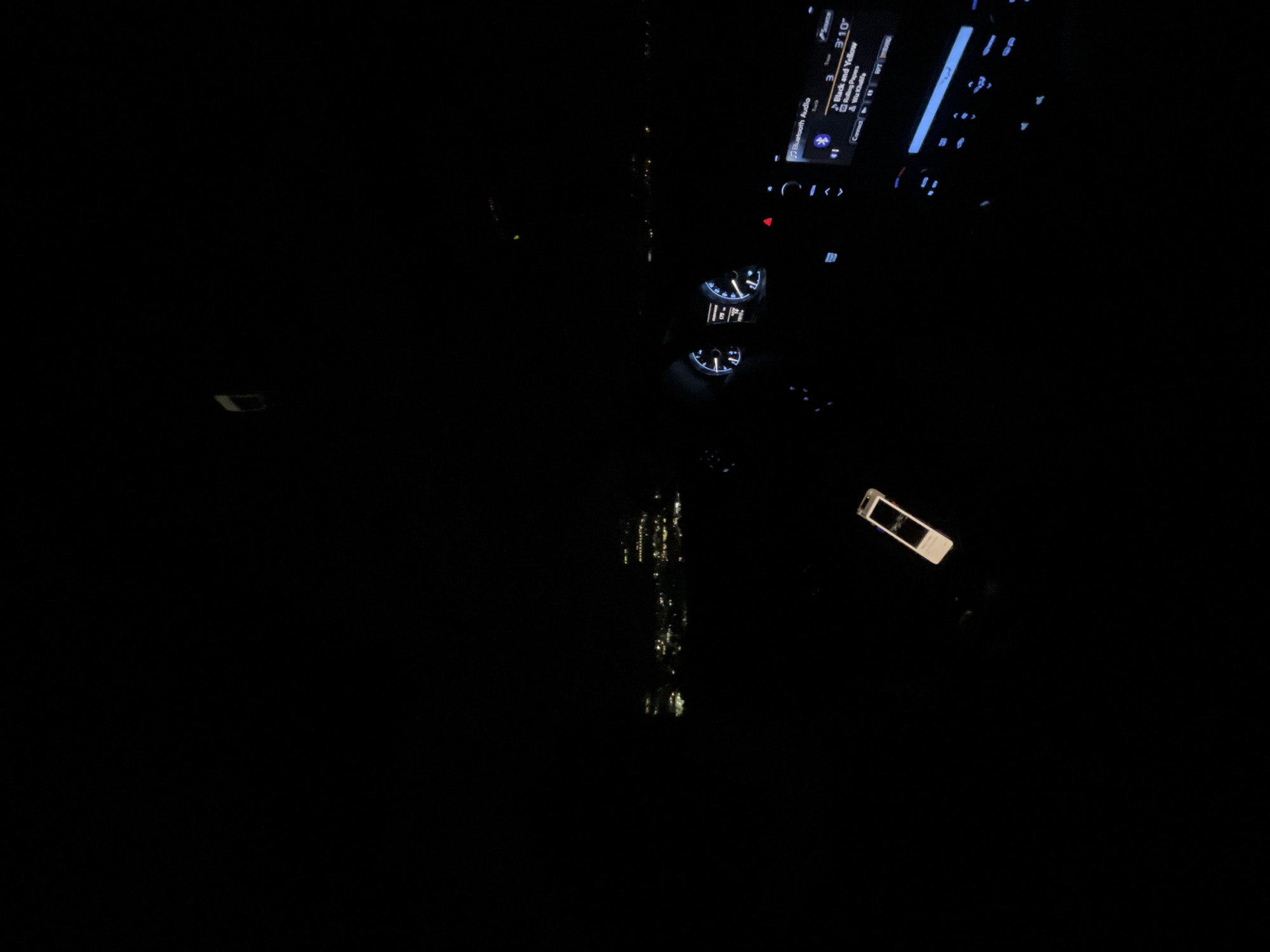 This image shows the interior of a car at night. The dashboard is visible, with illuminated gauges and a display screen showing that the song "Black and Yellow" by Wiz Khalifa is playing. Reflections of city lights can be seen through the windshield. There are no people visible. The location is unknown, but is said to be in Blagoevgrad, Bulgaria.