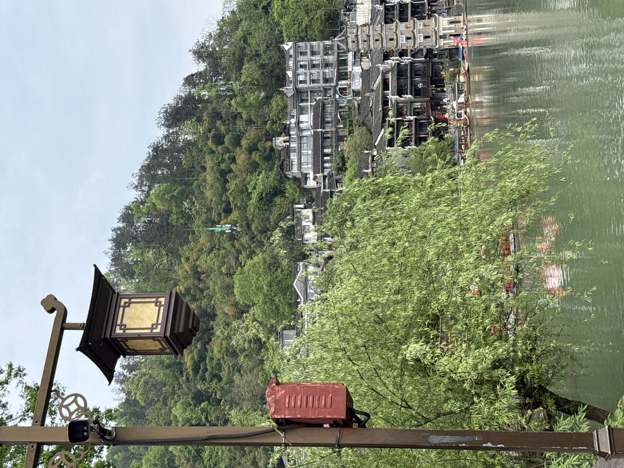 The image depicts a landscape in Fenghuang, China, showing a waterfront scene with traditional Chinese architecture. A hill densely covered in lush green trees slopes down to the water's edge. Several multi-story buildings with traditional tiled roofs and dark grey facades are built into the hillside, creating a stepped effect. The water is relatively calm, reflecting the buildings and trees. In the foreground, there is a traditional Chinese lantern with a dark brown frame and a light-colored translucent panel. Attached to the supporting post of the lantern is a security camera, and further down, a red box, likely housing electrical components, is affixed to the post. Part of the scene is obscured by green foliage, likely from a willow tree.