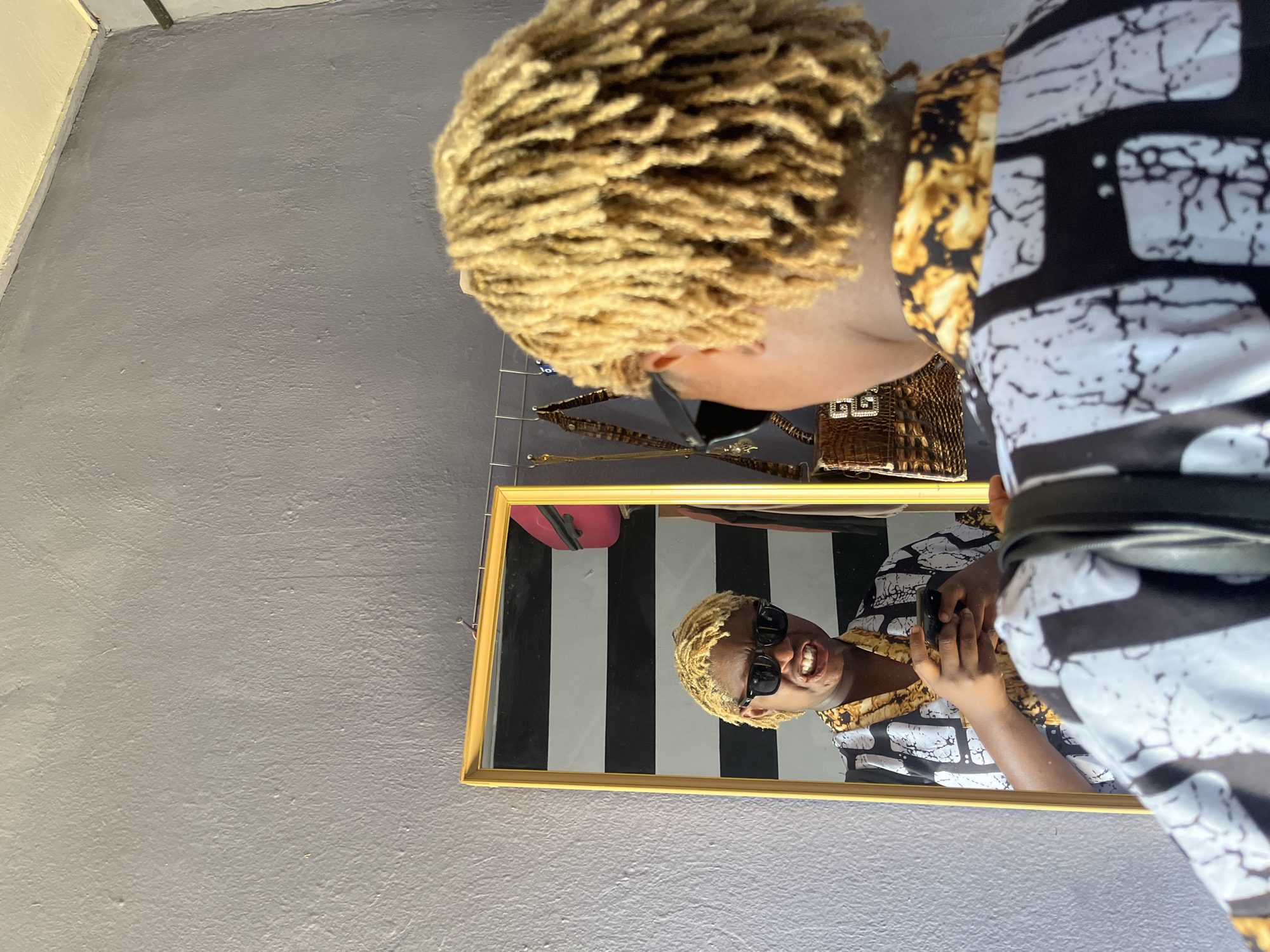 A person with blonde dreadlocks and sunglasses is taking a selfie in a room. The person is wearing a black and white patterned shirt with gold accents. In the reflection of a mirror, the person is holding a smartphone and making a grimace. Behind the person, there are various items hanging on a rack, including handbags and jewelry. The wall behind the mirror is painted a muted grey. The floor is not visible. The overall impression is that this is a personal space, likely a bedroom or dressing area, in a residence. The setting, specifically the style of the wall paint and the hanging rack, is consistent with homes found in urban areas of Nigeria, such as Jos.