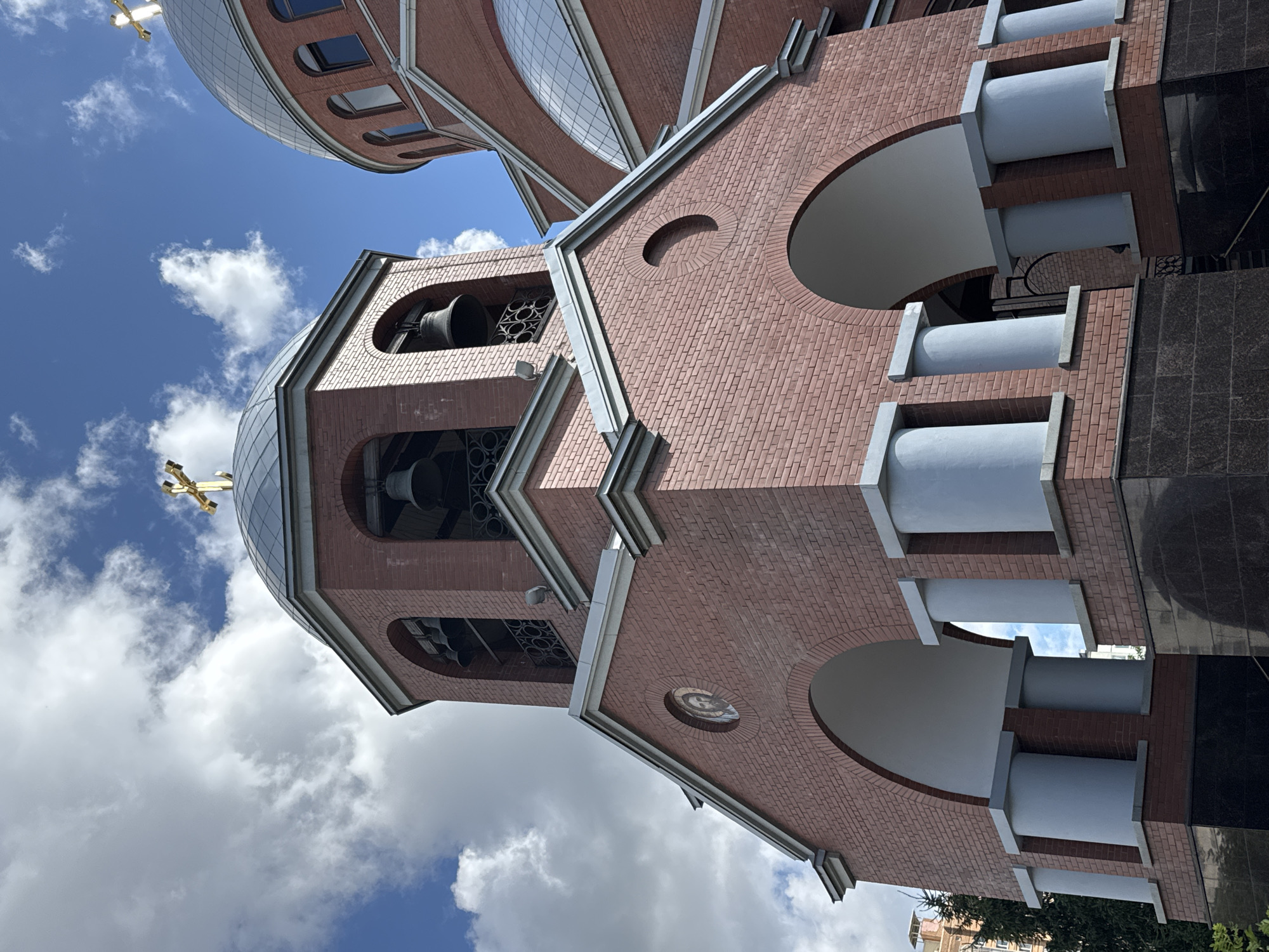 The image shows the exterior of a red brick church, likely in Saint Petersburg, Russia, under a cloudy blue sky. The foreground is dominated by the church's architecture, featuring a bell tower with arched openings revealing bells, and a domed roof topped with a golden cross. Further to the right, another dome and part of the church's facade are visible. Decorative elements include round arched windows and a circular mosaic depicting a religious figure on the front of the bell tower. The lower part of the structure is clad in dark polished stone. The overall scene is a detailed architectural shot of a religious building, emphasizing its construction materials and religious symbols.