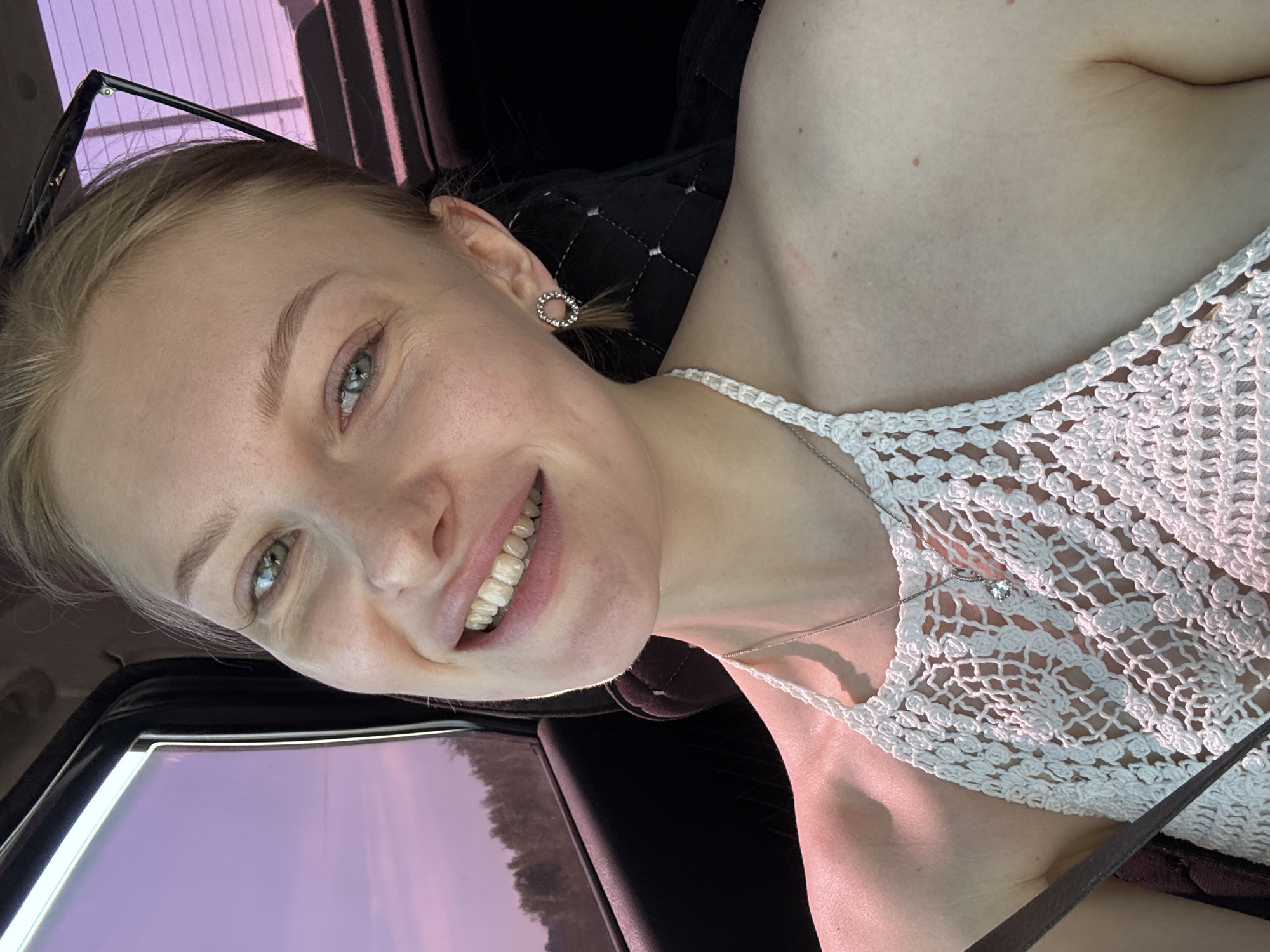 A fair-skinned woman with blonde hair and green eyes smiles towards the camera. She is wearing a white crochet top and a silver necklace with a heart-shaped pendant. Sunglasses are perched on her head. She appears to be sitting in a car, the interior is visible, and the window shows an obscured view of the outside. Based on the background through the car window, the location is not discernible and can not be determined as Perm, Russia, without additional visual cues or information.