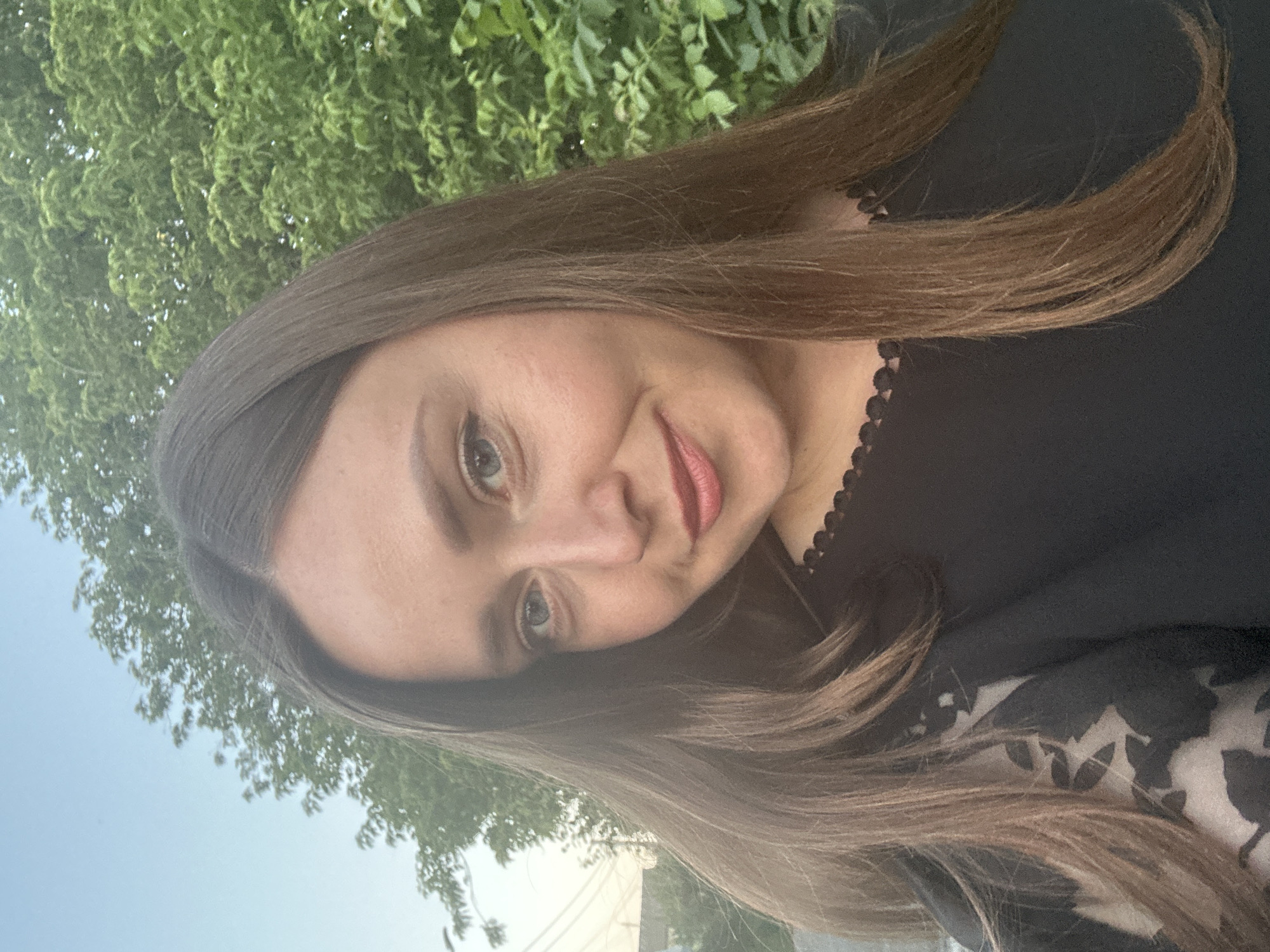 The image is a close-up, outdoor selfie of a woman with long, brown hair. She is wearing a black top with a scalloped neckline and a sheer, floral-patterned sleeve on her left side. The woman has light skin, blue eyes, and is smiling slightly. Her face is well-lit, and the background consists of green trees and a pale blue sky. The setting appears to be a residential area, possibly a park or backyard, with some power lines visible in the upper left corner, suggesting an urban or suburban environment. The specific location in Bila Tserkva, Ukraine, is not determinable from the visual information provided in the image.