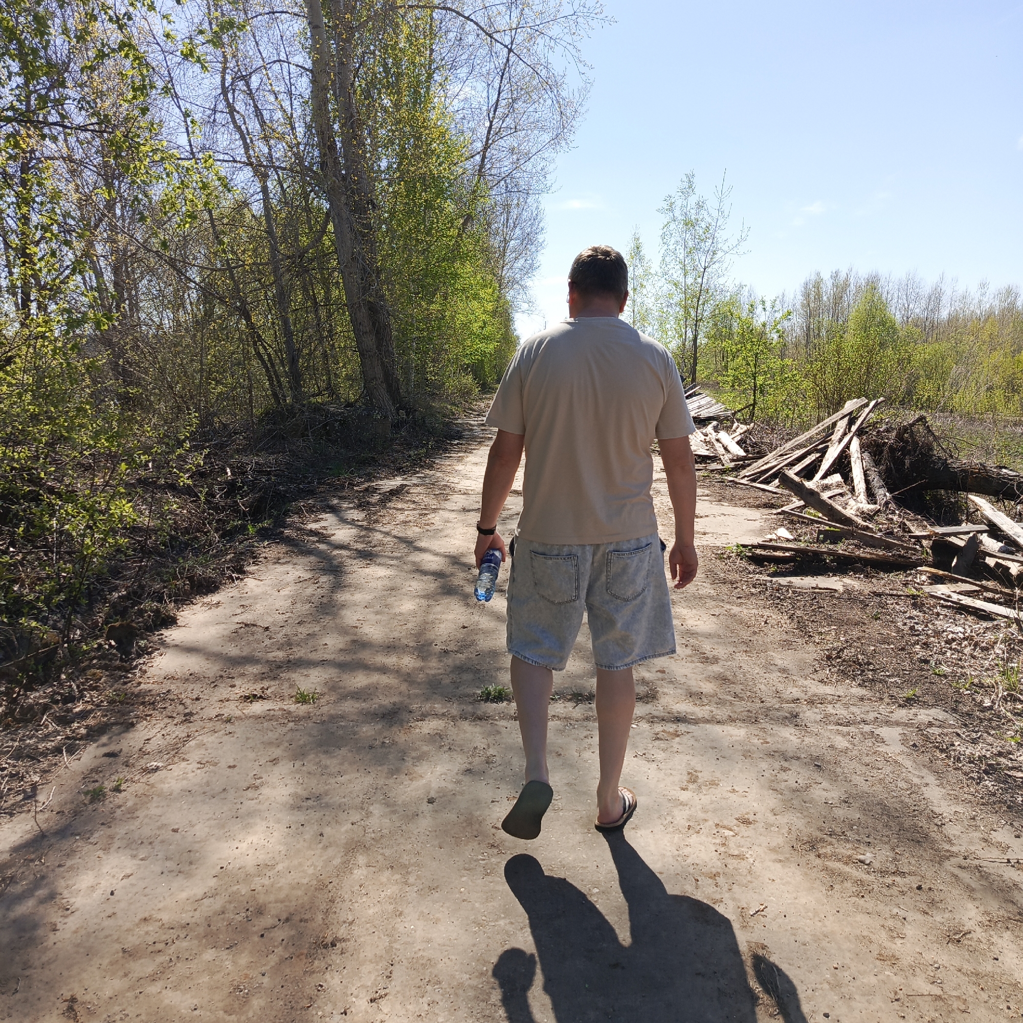 The image shows a man walking away from the camera on a dirt path. He is wearing a beige t-shirt, light denim shorts, and sandals. He is carrying a water bottle in his left hand. The path is surrounded by trees and shrubs, with some debris, including piles of wood, on the right side of the path. The sky is blue with a few clouds. The photograph was taken on a sunny day. The location is Chaikovsky, Russia.