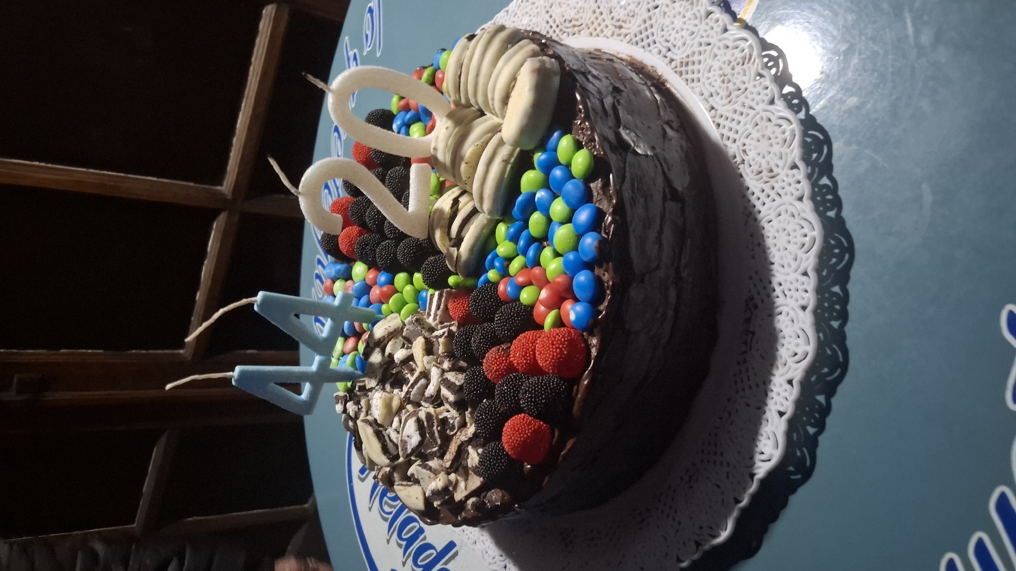 The image shows a chocolate cake, decorated with chocolate shavings, candy-coated chocolates (M&Ms), gummy candies resembling raspberries and blackberries, and white chocolate curls. White number "20" candles and a blue number "4" candle are inserted into the cake. The cake is placed on a white doily and is sitting on a blue surface with writing in white. The location is likely Buenos Aires, Argentina, based on the writing on the blue surface, although the writing is not entirely visible to confirm the location. The cake is placed near a set of shelves in a dimly lit room.