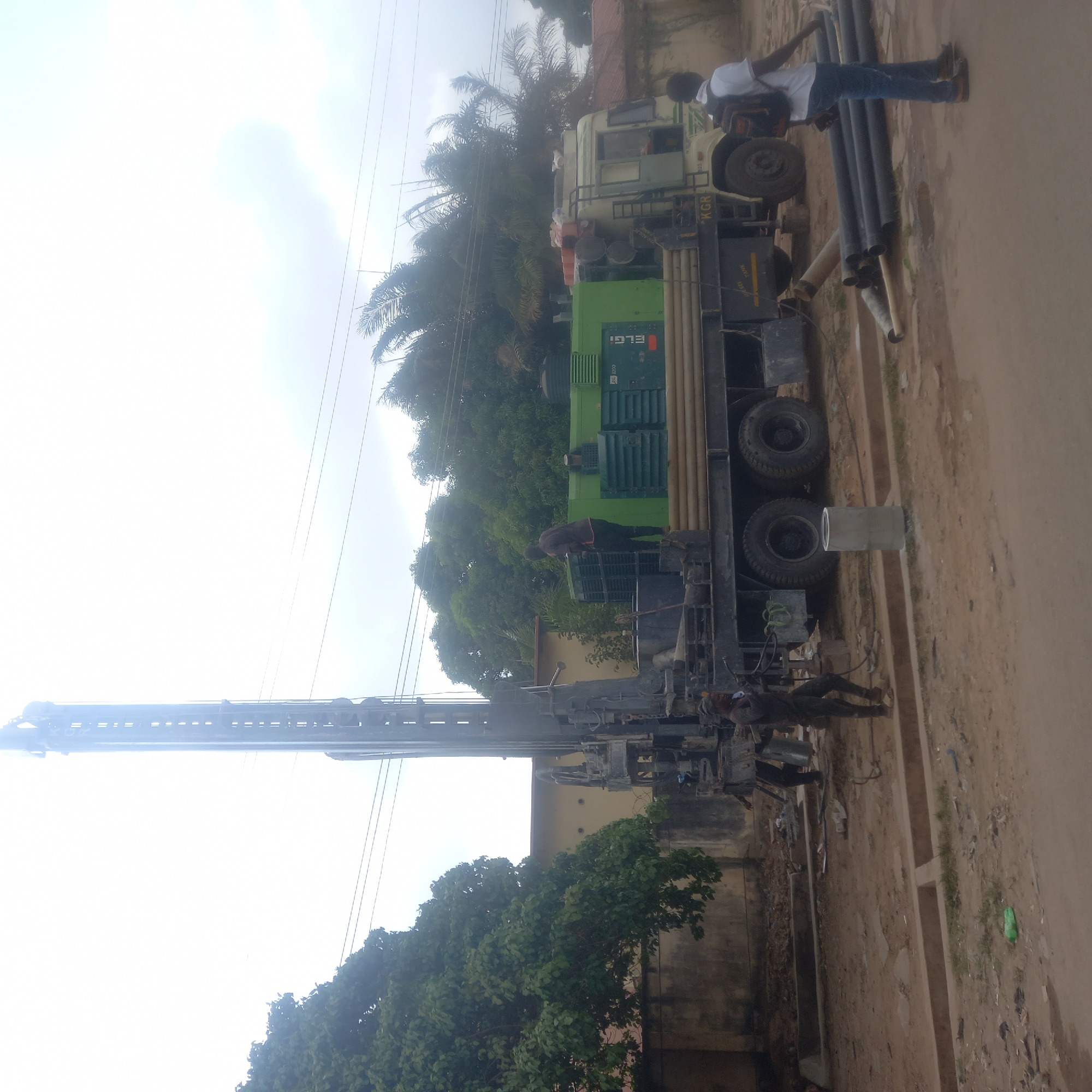 This media file captures a borewell drilling operation taking place during the daytime in Dadi Riba, Nigeria. The central subject is a large, truck-mounted drilling rig with a tall, extended mast, suggesting it is either actively drilling or being set up/dismantled.
Several individuals are present, appearing to be workers involved in the drilling process. One man, dressed in a white shirt and blue jeans, stands next to a stack of black pipes (likely casings) on the ground. Another worker in dark clothing is visible near the green generator unit on the rig, and more workers are scattered around the machinery.
The drilling rig itself is complex, featuring a white truck cab and a distinctive green generator unit clearly labeled "ELGI". The side of the rig's frame also shows text including "KGR" and some partially legible numbers like "KM 120" and "TEST". A cylindrical object, possibly a section of casing or a support, rests on the dirt ground near the truck's wheels.
The setting is outdoors on what appears to be a dirt road or open ground, flanked by lush green trees, including tall palm trees, in the background. Partial views of buildings are visible behind the foliage. The sky is bright, indicating daylight, though it appears somewhat hazy or overexposed in parts. The ground is dry and dusty. The overall scene depicts industrial activity related to water infrastructure development in a rural or semi-urban environment.