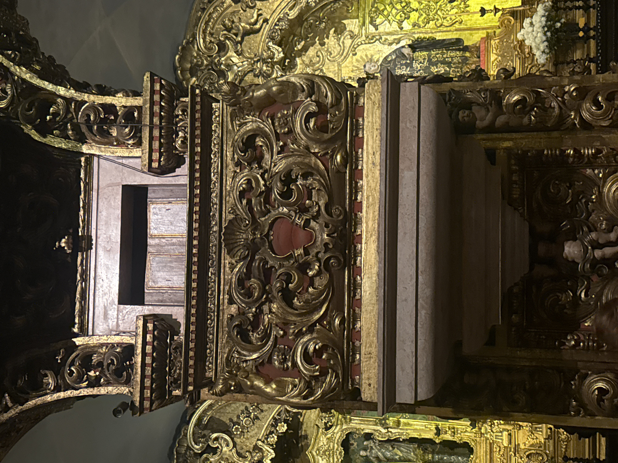 The image shows a detailed view of ornate golden baroque architecture, likely within a church or religious building in Rio de Janeiro, Brazil. The foreground features a substantial, intricately carved golden structure with red accents, reminiscent of an altar or a pulpit. This structure is adorned with swirling decorative elements, cartouches, and small sculpted figures of cherubs or putti. To the left and right, the background reveals more gilded religious art, including statues of figures in traditional attire. One statue on the right appears to be a cleric or saint. White flowers are visible in a vase on the lower right, adding a touch of natural beauty to the opulent setting. The lighting is dim, highlighting the golden surfaces and casting shadows that emphasize the elaborate carvings. The overall impression is one of historical grandeur and religious devotion.