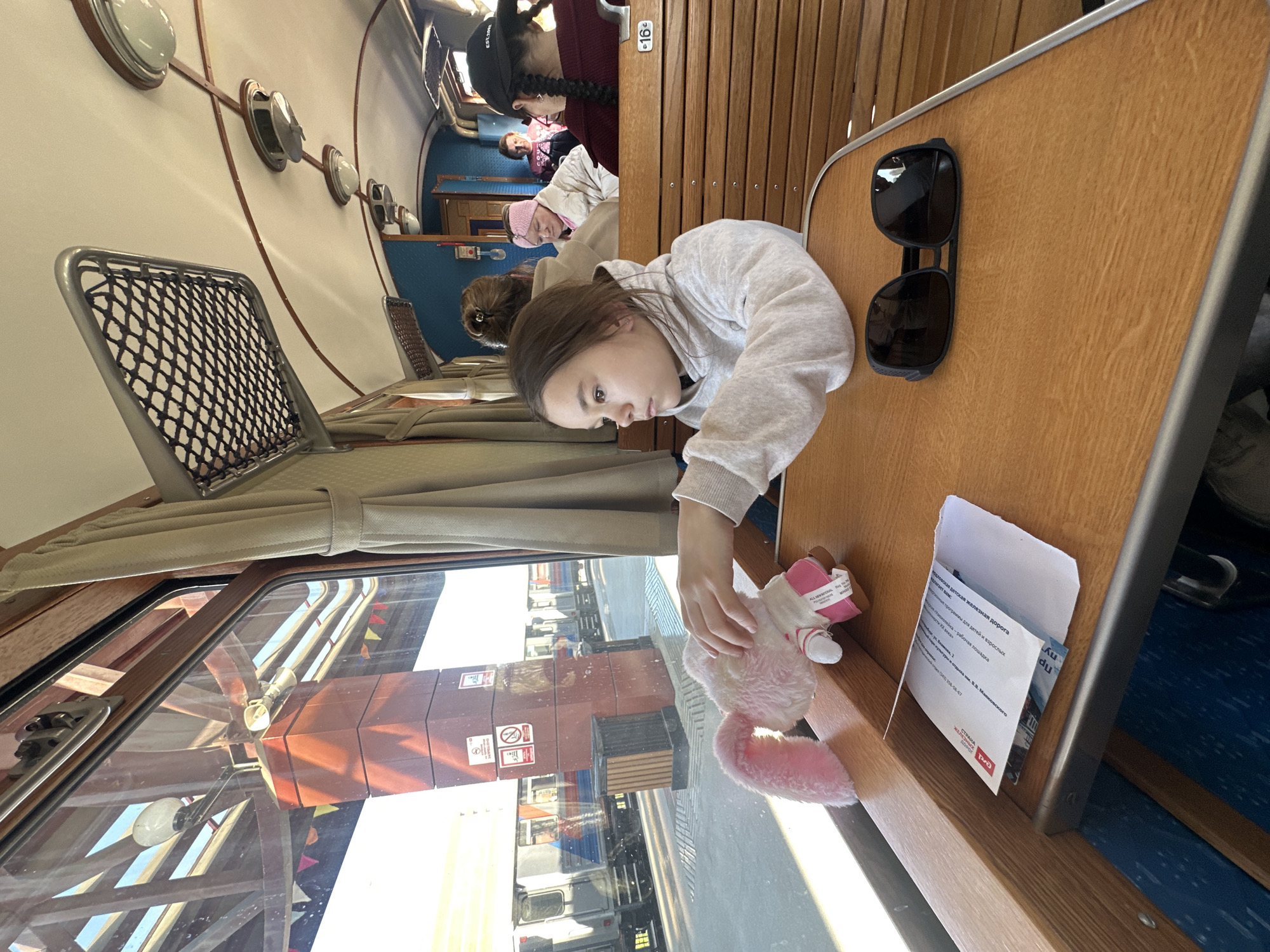 A young girl with light brown hair, wearing a gray hooded sweatshirt, sits at a wooden table on a train. She is looking at and touching a pink stuffed rabbit. Sunglasses and a folded informational pamphlet, written in Russian, lie on the table near her. The text refers to the "Frolkovskoy Children's Railway." Outside the window, a blurred view of an urban environment is visible with signs and commercial vehicles, reflecting the light. Other passengers are seated nearby in the train carriage. The number "16" is visible on the back of a seat ahead.