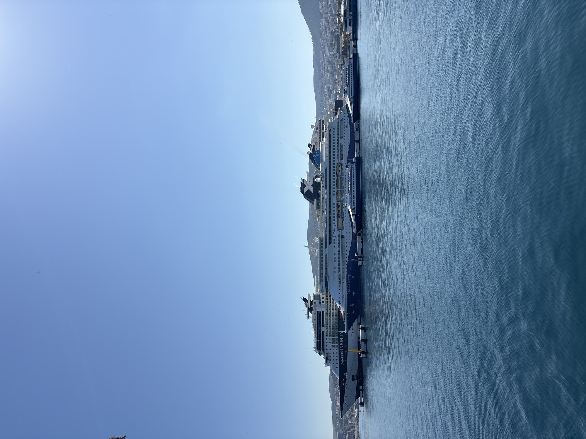 The image displays a bustling port scene in Kuşadası, Turkey, under a clear and bright blue sky, suggesting a sunny day. Several large cruise ships are docked along a pier, occupying the midground of the frame.
The ships are predominantly white and navy blue, featuring multiple decks, lifeboats, and antenna structures on their upper levels. The name "CELEBRITY INFINITY" is clearly visible in white lettering on the dark blue hull of the ship closest to the viewer. Behind it, another similar cruise ship is docked, with "CELEBRITY C..." and an 'E' visible, indicating it is likely another Celebrity Cruises vessel (possibly "Celebrity Equinox" or "Celebrity Eclipse"). The calm, deep blue water in the foreground and between the ships shows subtle ripples, reflecting the clear sky.
In the background, a coastal town extends along the shore, with buildings nestled against the foothills of prominent, green-brown mountains or hills. The sky is a vast, unobstructed expanse of light blue, with a bright sun creating a soft glare in the upper left corner of the frame. No specific human figures or activities are discernable due to the distance. The overall scene conveys a sense of a vibrant tourist port during good weather.