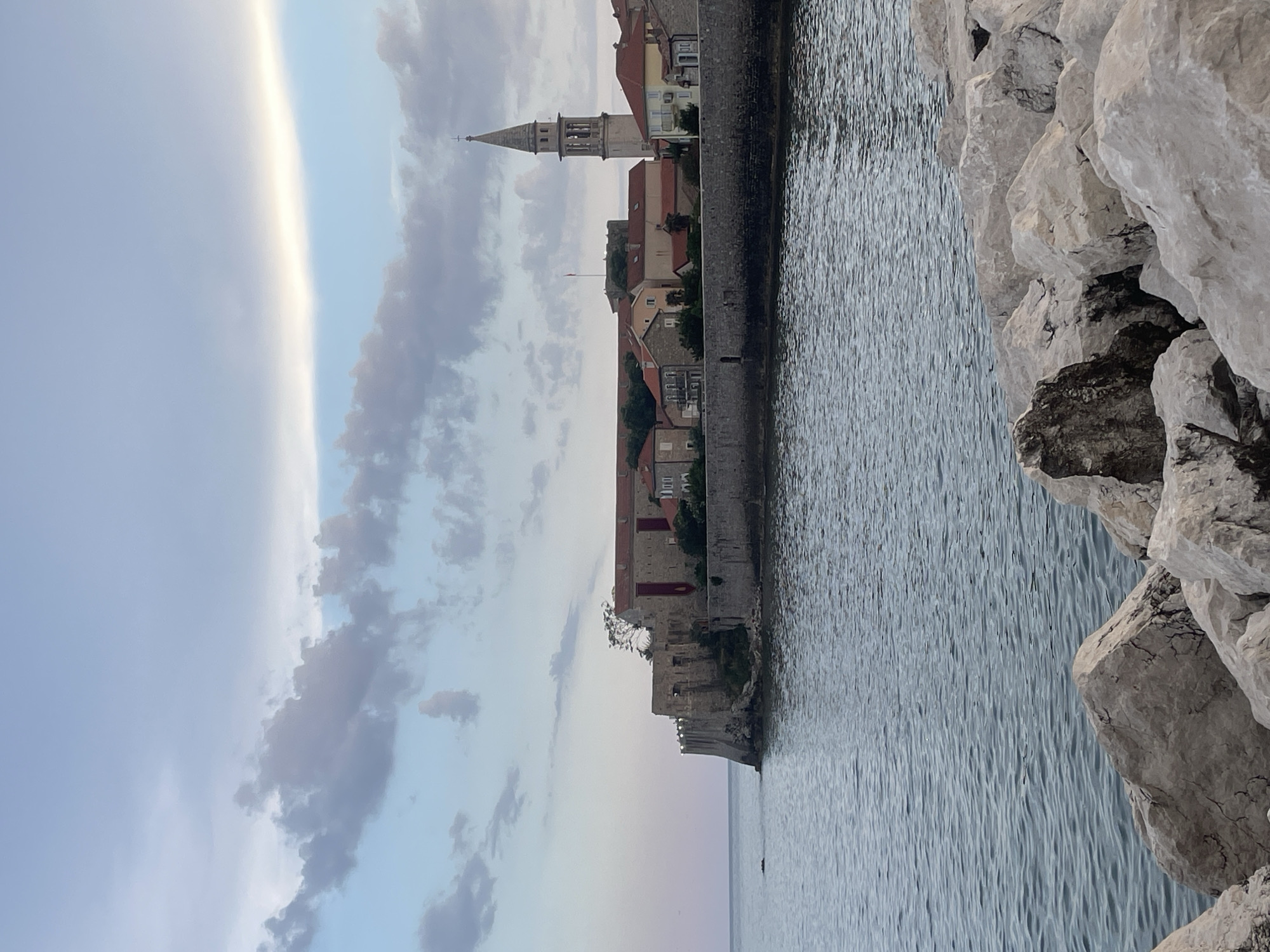 The image shows a coastal view of a fortified town. In the foreground, large, rough-hewn rocks form a barrier along the edge of the water. The sea is a deep blue-grey, with a slightly rippled surface. Beyond the water, a stone wall encloses a section of the town, which features buildings with red-tiled roofs. A tall, slender bell tower, characteristic of a church, rises above the other structures. The sky above is a soft blue, with scattered clouds in muted tones of grey and white. The overall impression is of an old European coastal settlement, possibly at dusk due to the soft lighting and subdued colors.