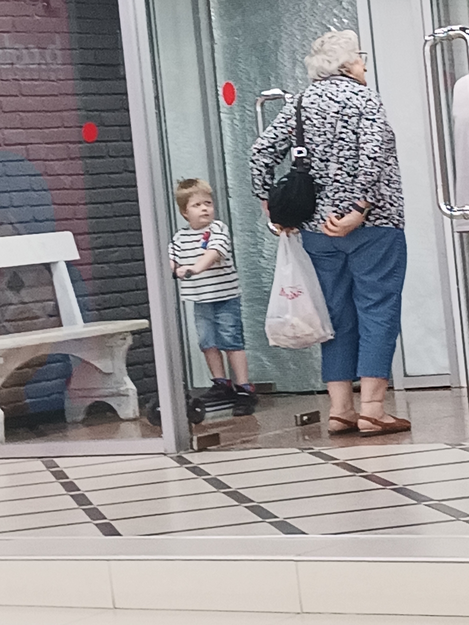 A young boy with blond hair stands on a black scooter, facing forward. He wears a striped t-shirt and denim shorts. Behind him, an older woman with gray hair and glasses stands with her back to the camera. She has a black bag slung over her shoulder and is holding a white plastic bag in her right hand. She wears a patterned jacket and blue capri pants. The scene appears to be indoors, possibly a public building or mall, with a tiled floor and a glass door. The background includes a brick wall and a white bench. There are no visible landmarks that would specifically place this image in Chișinău, Moldova.