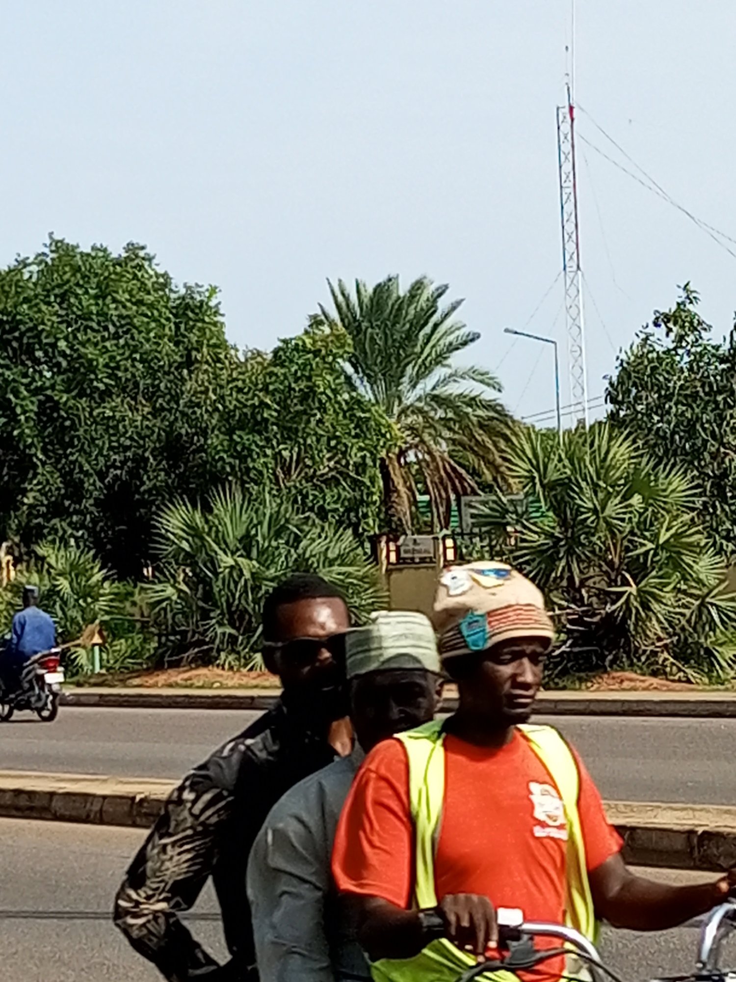 In Garko, Nigeria, three men are riding on a motorcycle in the foreground, with a fourth man on a separate motorcycle in the background. The man in the front is wearing an orange t-shirt and a high-visibility yellow vest, along with a striped cap. The man behind him is wearing a dark patterned shirt and sunglasses, and the third man is wearing a light-colored hat. The background features lush green trees and palm trees, a street light, and a tall transmission tower with a red marking at its top. The sky is clear and pale blue.