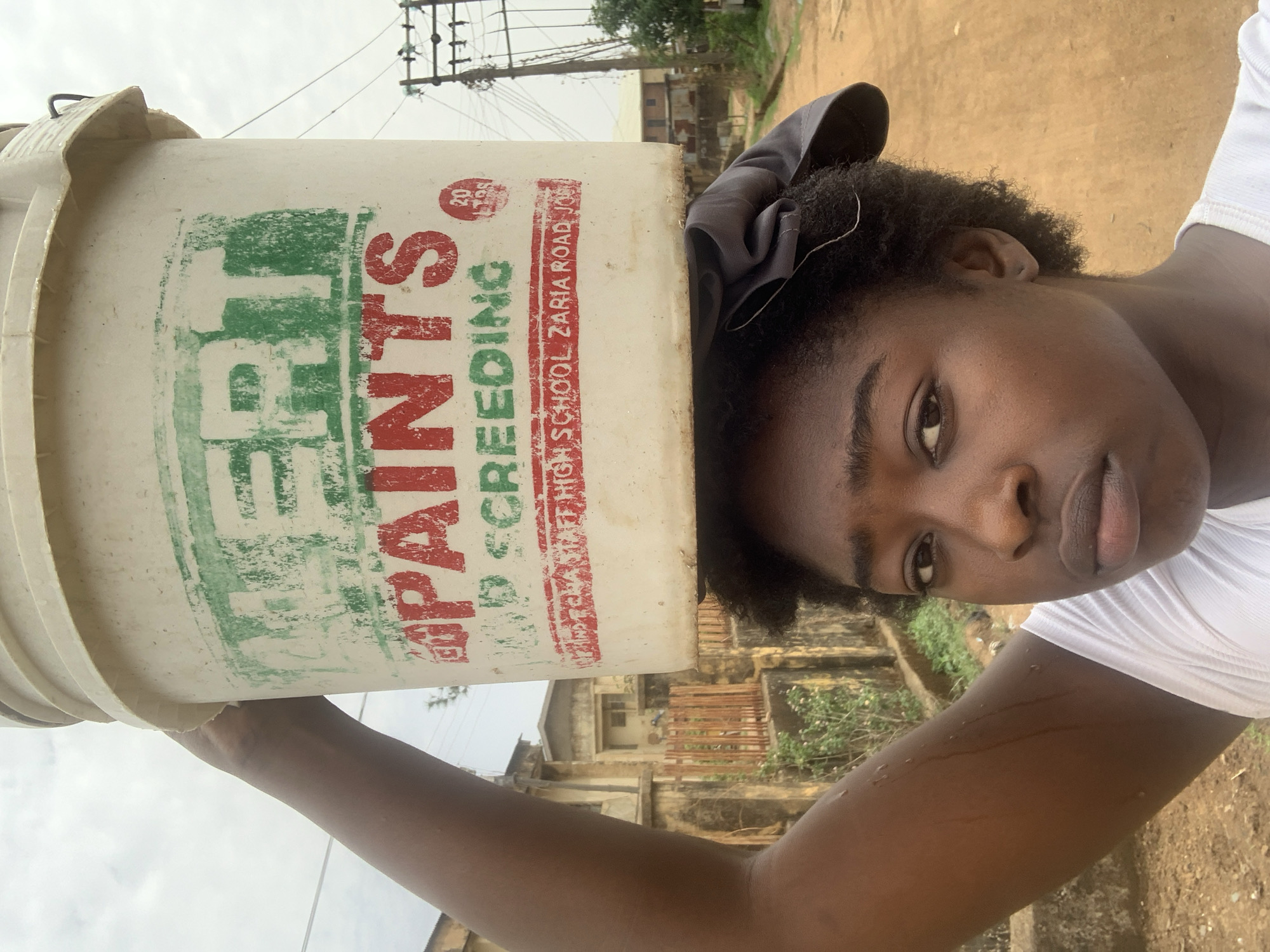 A young woman is holding a white bucket with the inscription "ERT Paints Screeding High School Zaria Road Jos" on it. The location is Rafin Par, Nigeria. The background includes a building and a utility pole. The woman is wearing a white shirt and a hair bow.