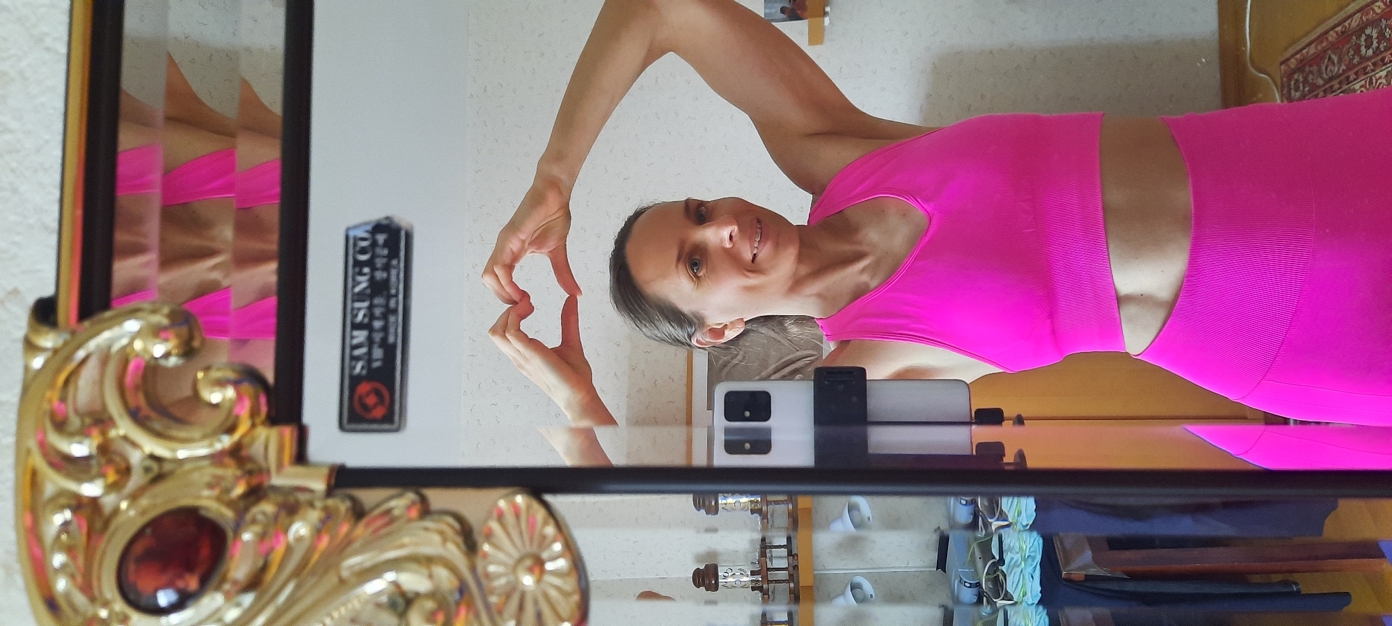 The image is a mirror selfie taken indoors, likely in a home in Moscow, Russia. It features a woman, appearing to be in her 30s or 40s, with light hair pulled back and fair skin. She is smiling cheerfully at her reflection while making a heart shape with her hands held near her face. She is dressed in a vibrant, hot pink athletic two-piece set, comprising a sleeveless crop top and high-waisted leggings that expose a sliver of her midriff.
Her smartphone, used to capture the image, is visible resting vertically on a surface directly below her chest, with its light-colored case and dark camera module facing away from the mirror. The mirror itself is partially framed on the left by an ornate, gold-colored decorative frame featuring a dark red or brown jewel-like embellishment.
In the reflection, to the left of the woman's head, a rectangular black object is visible, seemingly mounted on a wall. It bears white text that clearly reads "SAM SUNG CO." followed by Korean characters and "MADE IN KOREA". The background walls are light-colored, possibly with a subtle texture or pattern. A hint of a patterned rug or carpet is visible on the right side of the image, behind the woman's legs. The scene is brightly lit, suggesting it is daytime. The woman's smile and hand gesture convey a positive and friendly emotion.