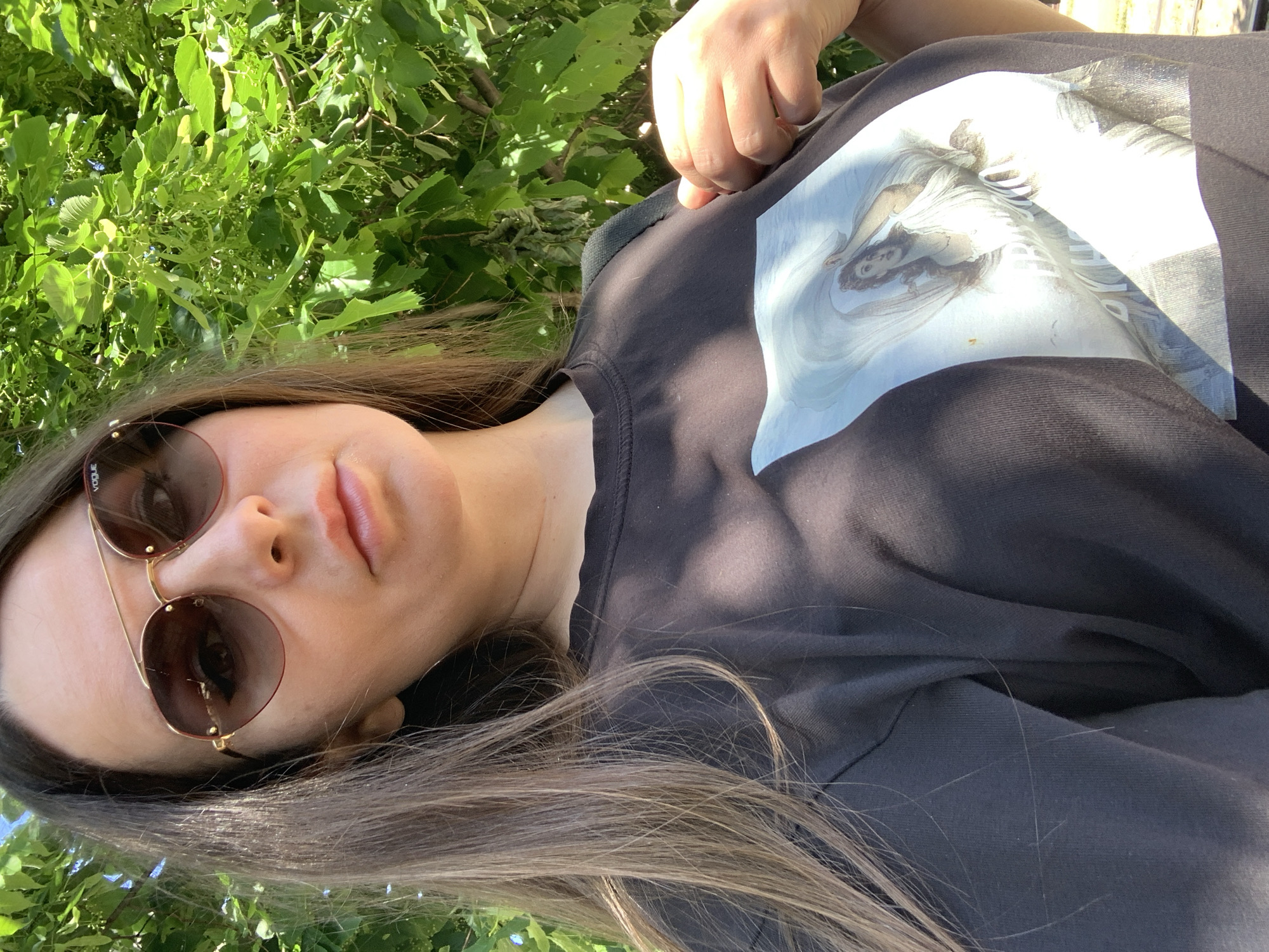 A close-up, rotated portrait shot captures a person, likely a woman, reclining outdoors amidst lush green foliage. The subject is seen from a slightly overhead perspective, looking upwards towards the camera with a neutral to relaxed expression. Her long, light brown hair spreads out around her head. She is wearing round, gold-rimmed sunglasses, with the brand name "VOGUE" subtly visible on the left lens. Her lips are slightly parted.
She is dressed in a dark-colored, possibly black or dark grey, t-shirt or sweatshirt. A prominent graphic print on the front of her top depicts a stylized image of a woman's face and upper body, rendered in light tones against the dark fabric. The image resembles classical art or an angelic figure with flowing hair and drapery.
The background is dominated by vibrant green leaves and branches, suggesting a natural outdoor setting such as a park or garden. Bright sunlight illuminates the scene, creating strong highlights on the foliage and the subject's face and clothing, indicating a clear, sunny day. Shadows are cast by the surrounding leaves onto the person's shirt. The image's orientation suggests the person is lying down, with the camera positioned above them.