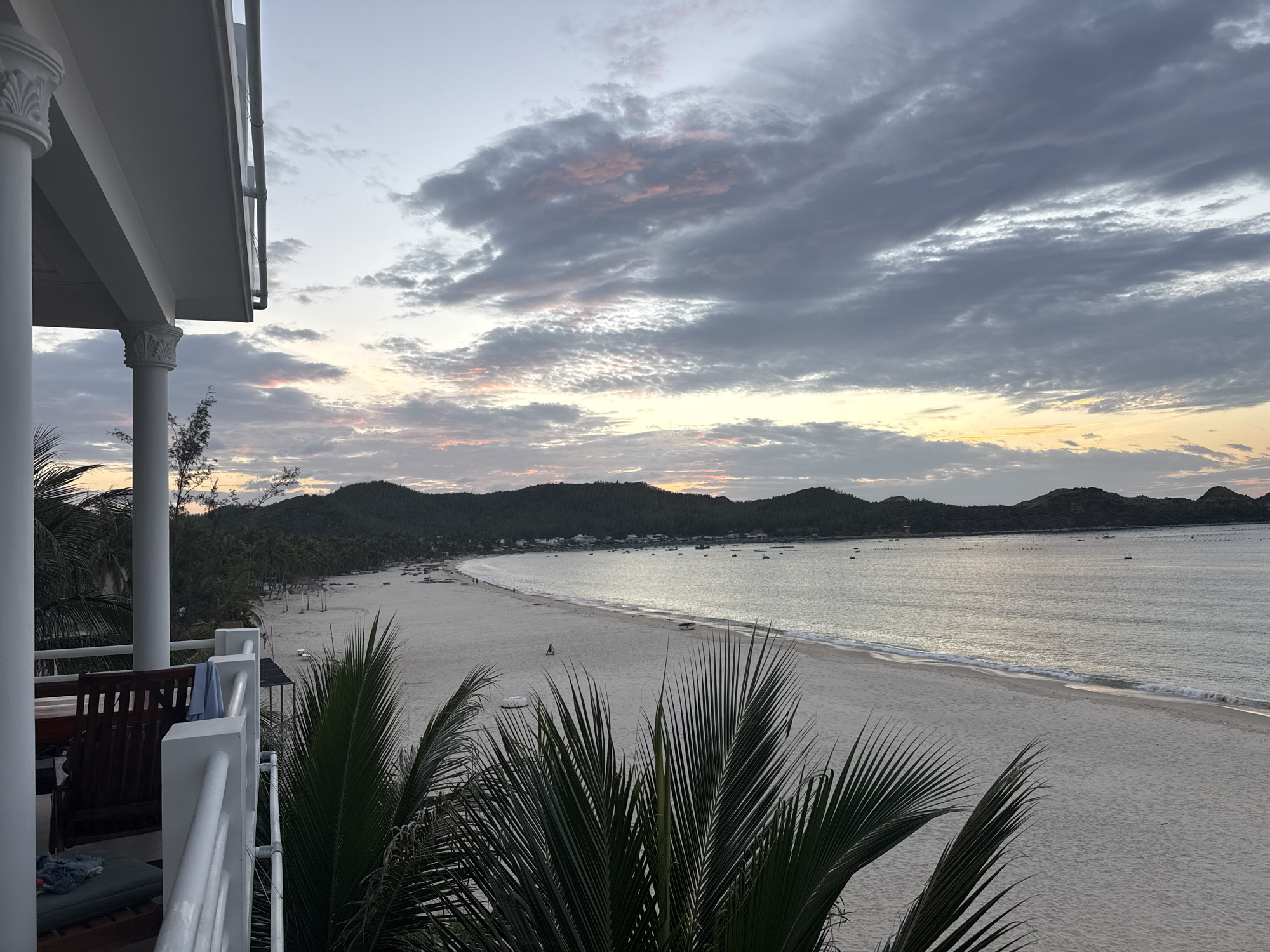The image depicts a serene beach scene in Song Cau, Vietnam, during what appears to be early morning or late evening, as indicated by the soft, diffused light and the colors in the sky. The foreground is dominated by lush green palm fronds, framing a view of a wide, sandy beach that stretches along a calm body of water. In the distance, a line of rolling hills covered in dense vegetation forms the horizon.
On the left side of the frame, a portion of a white building with elegant columns and a balcony railing is visible, suggesting a resort or a private residence overlooking the beach. A dark wooden bench is placed on the balcony.
The beach itself is largely empty, with a few scattered figures walking along the shore. Several small boats are anchored in the water, and what appear to be fishing nets or buoys are visible further out at sea. The water is tranquil, with gentle waves lapping at the shore. The sky is a canvas of soft grays, whites, and hints of pink and orange, indicative of either sunrise or sunset, with scattered clouds adding texture and depth to the scene. The overall impression is one of peace and natural beauty.