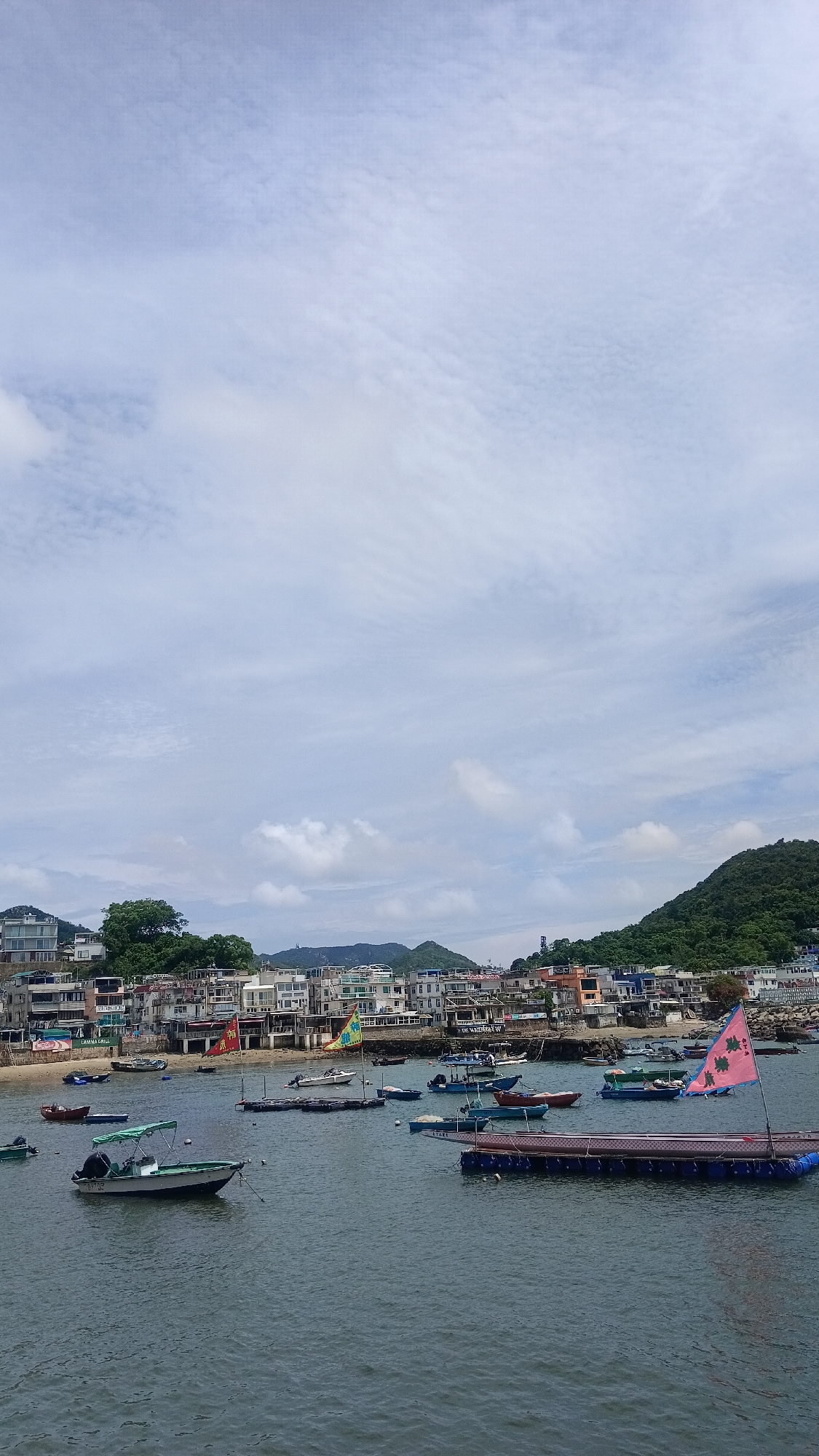 This is a landscape photograph of a coastal village in Hong Kong. The image depicts a harbor filled with numerous small boats of various sizes and designs, some moored and others seemingly at anchor. In the background, a densely populated village lines the shore, with buildings of differing heights and architectural styles clustered together. Beyond the village, lush green hills rise, indicating a hilly or mountainous terrain. The sky is partly cloudy, with a mix of white clouds against a pale blue backdrop. The overall scene suggests a tranquil day in a seaside community. The presence of the flag on one of the boats with Chinese characters confirms its location in Hong Kong. The image appears to have been taken from a slightly elevated vantage point, possibly from another boat or a pier.