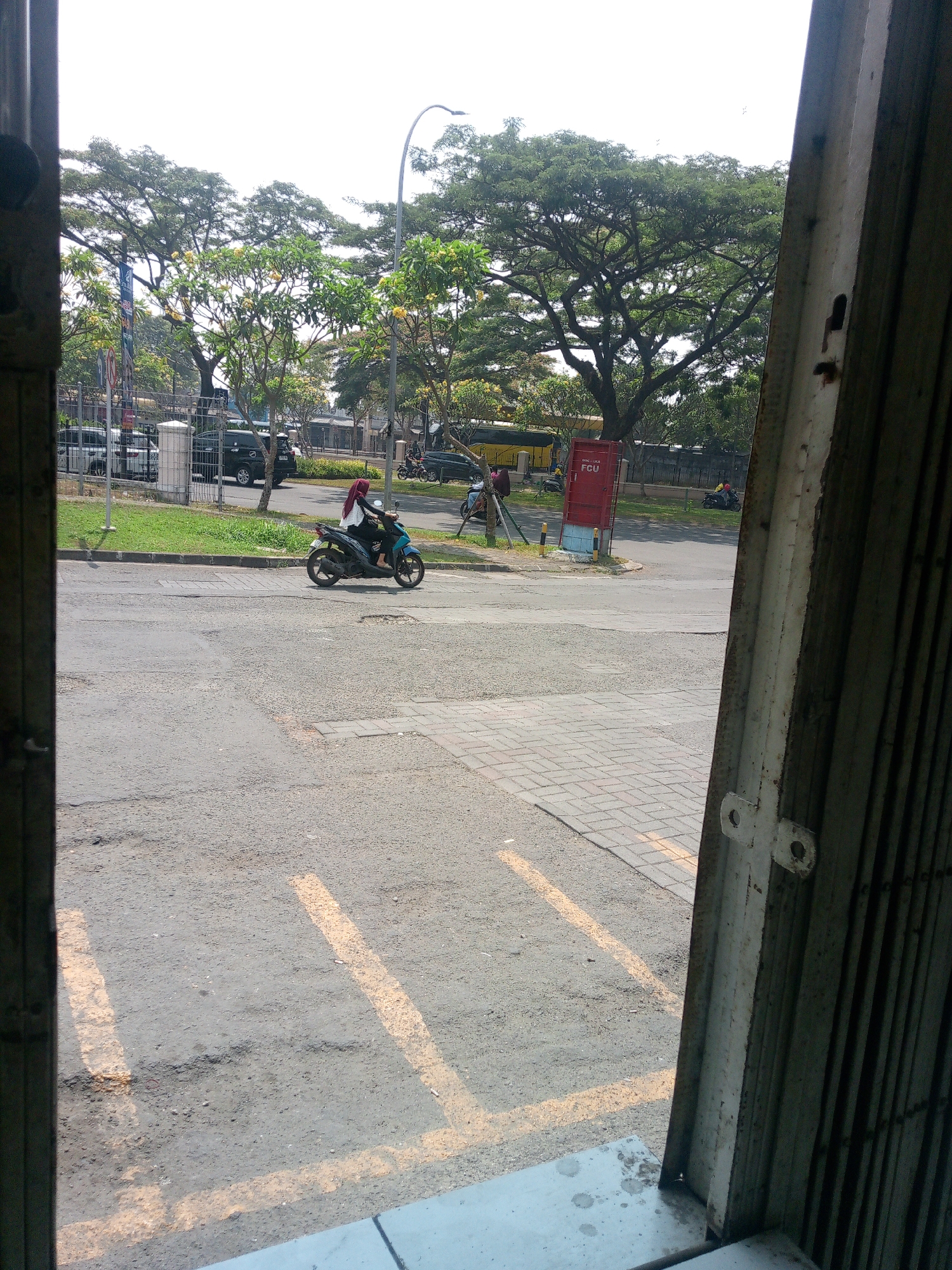 The image depicts an outdoor scene with a paved road and sidewalk in the foreground. A woman wearing a hijab rides a blue and black motorcycle on the road, facing towards the right of the frame. Behind the motorcycle, a large, mature tree with dense foliage dominates the background, with other trees and buildings visible further beyond. To the right, a rusty metal gate or door is partially visible, framing the scene. The environment appears to be a street in Cikupa, Indonesia, characterized by urban elements like a road, sidewalk, and parked vehicles, as well as natural elements like trees. The time of day seems to be daytime, indicated by the bright lighting and shadows. There is also a red utility box with "FCU" written on it.