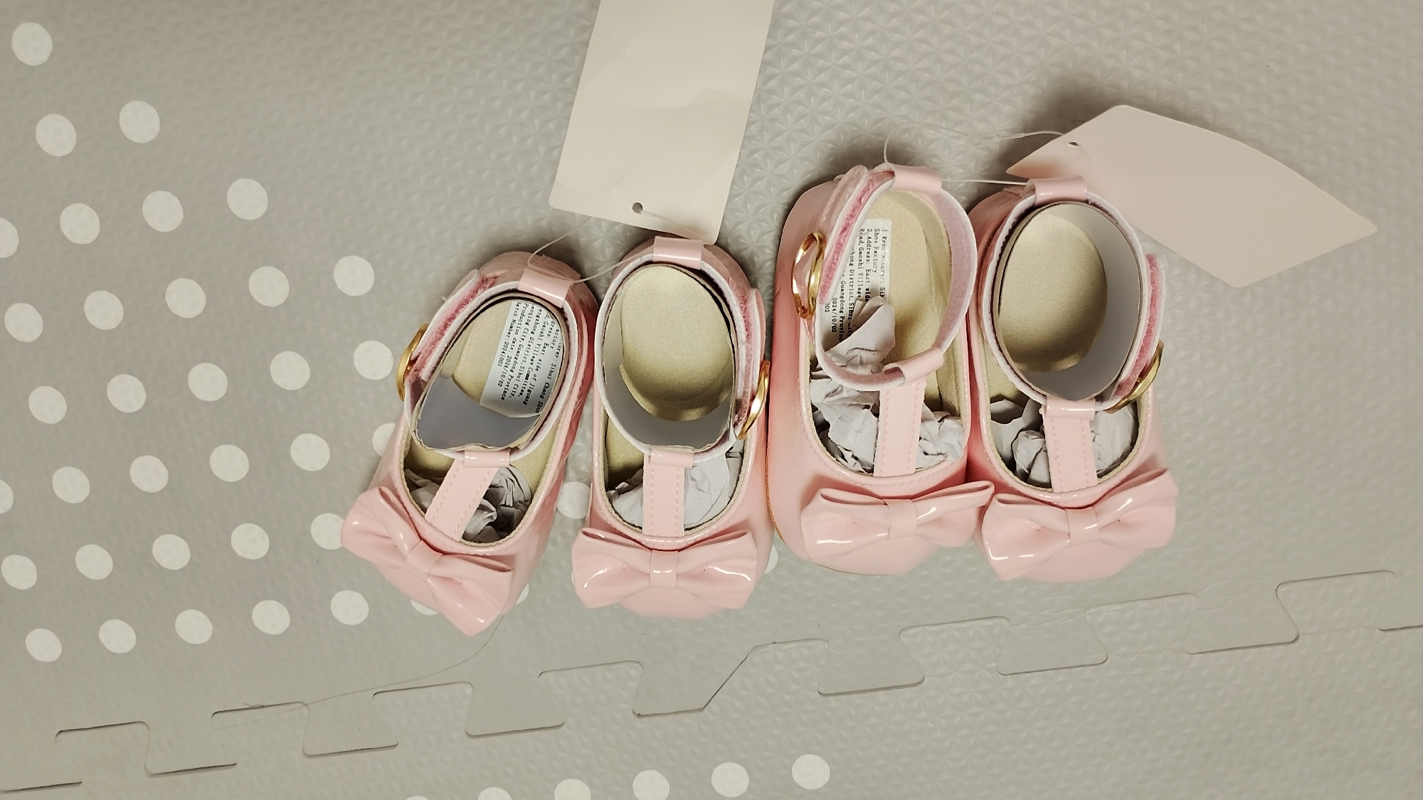 The image shows a pair of pink baby shoes with bows, laying on a grey mat with white polka dots. There are two blank tags attached to the shoes. The scene is indoors, likely in a home environment. The image does not contain any information about the city of Smolyan, Bulgaria.