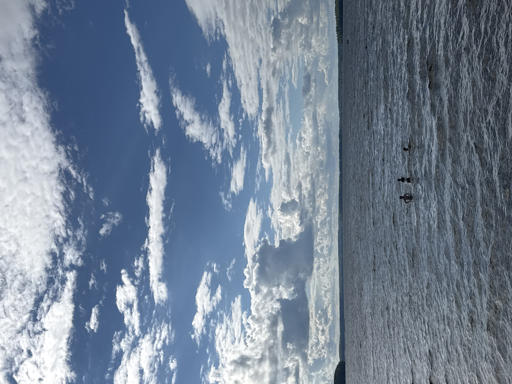 This image depicts a scenic view of a body of water under a partly cloudy sky. The water appears to be a lake or a wide river, with visible ripples and waves suggesting a gentle breeze. Several people are present in the water, some appearing to be swimming or wading. One person is captured mid-jump, adding a dynamic element to the scene. In the distance, a tree line marks the edge of the land, with a hint of a structure or boat visible near the shore.
The sky is a vibrant blue, interspersed with fluffy white clouds, indicating a pleasant, possibly sunny day. The lighting suggests it could be daytime, likely afternoon given the sun's position and the overall brightness. The overall mood conveyed is one of leisure and enjoyment of the outdoors. The location context provided is Desnogorsk, Russia, which suggests this is likely a natural body of water near the city. There is no visible text in the image.