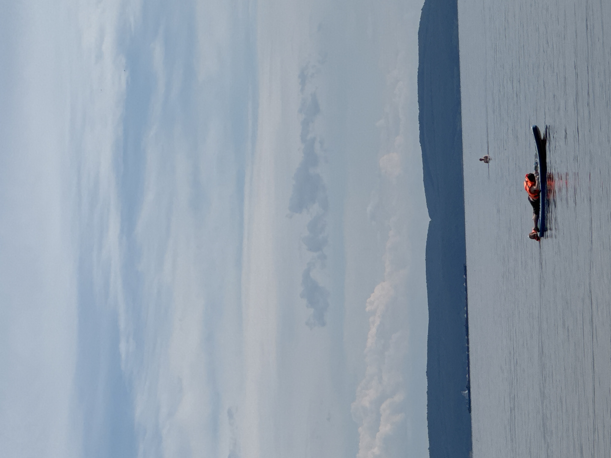 A person wearing an orange life vest is lying prone on a blue paddleboard in a calm body of water. The paddleboard is positioned in the foreground, slightly to the left of center. In the mid-ground, another person is visible standing in the water, further out from the shore. In the background, a long barge or ship with a series of connected smaller vessels is moving across the water, and a dark, tree-covered hill forms the horizon line. The sky above is cloudy with a mix of gray and white clouds. The scene appears to be a recreational activity on a lake or large river.