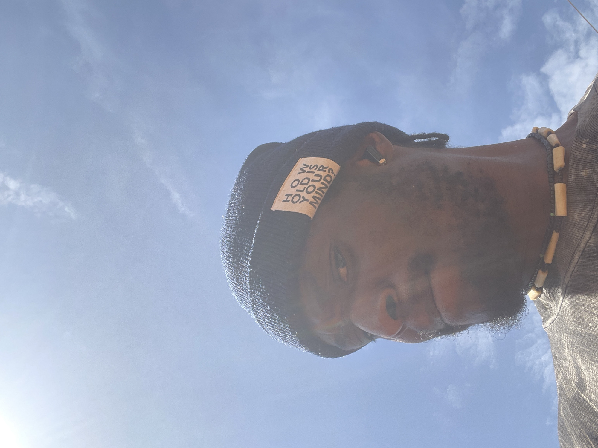 The image is taken from a low angle, looking up at a person against a blue sky with scattered clouds. The person appears to be a black man wearing a dark beanie with a rectangular label featuring the words "HOW DO YOU MIND?" written vertically. He has a short beard and wears a necklace consisting of dark beads interspersed with cylindrical, light-colored beads. An earbud is visible in his right ear. The overall lighting suggests bright daylight. The specific location within the city of Jos, Nigeria, is not discernible from the image.