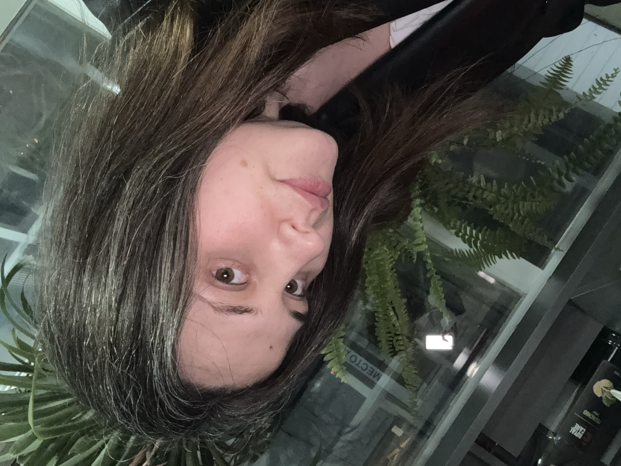 A rotated medium shot depicts a woman with dark brown hair, fair skin, and brown eyes. She faces the camera with a slight tilt of her head. She wears what appears to be a black jacket or top over a white garment. The background shows indoor foliage including ferns adjacent to the woman's face on the left and right. Further behind, a window reflects light and vegetation. Text in Cyrillic script is visible on a sign within the window reflection. The bottom right of the image contains a dark bottle labeled "ROYAL CANE PISTACHIO" in English. Given the Cyrillic script, this scene is likely taken in a country that uses that alphabet. While I can tell you that the script visible on the sign within the window reflection is Cyrillic, I can't verify the location in the city of Chaikovsky, Russia.