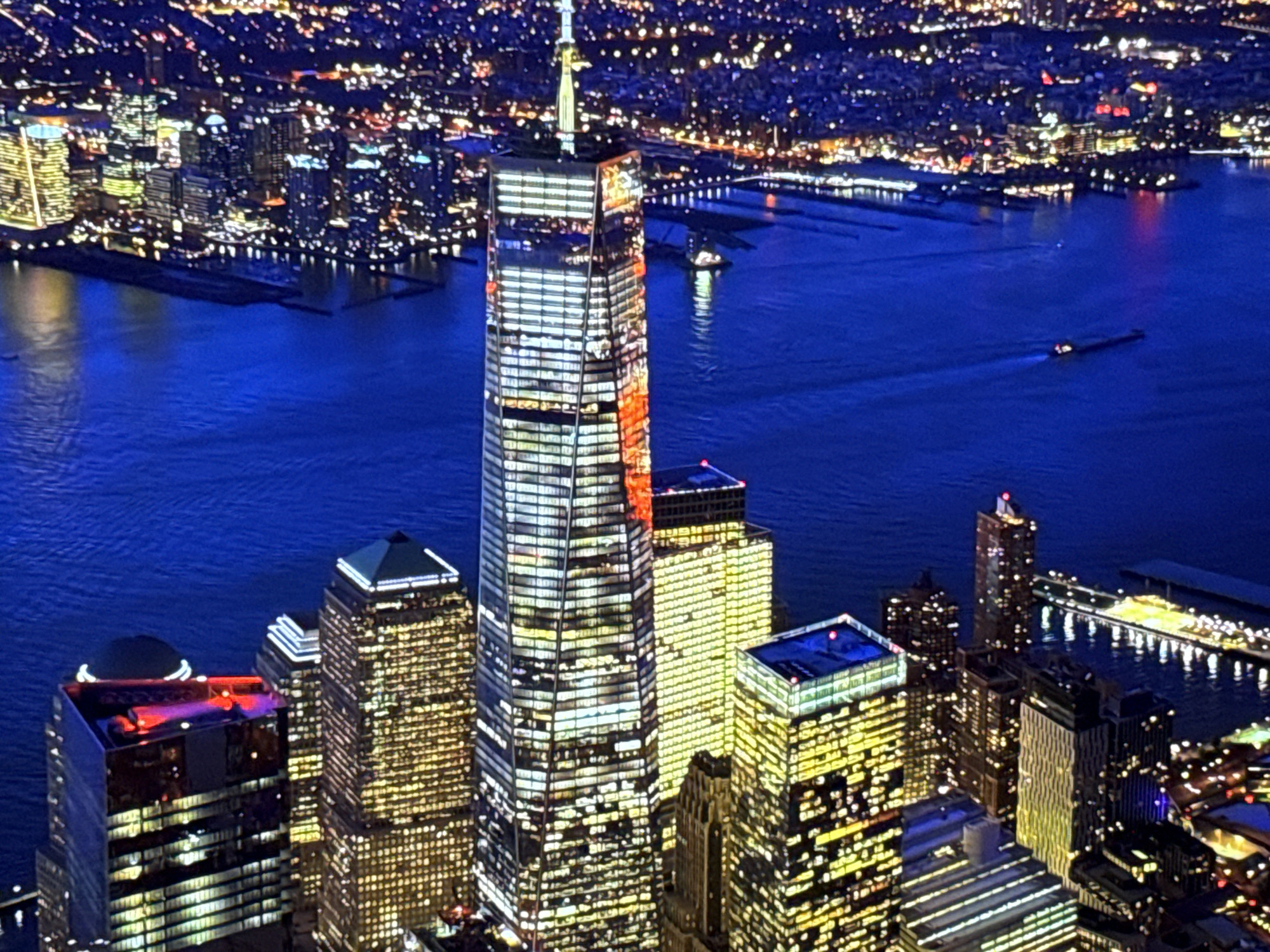 This is a nighttime aerial photograph of a densely built urban area with numerous skyscrapers. The primary focus is the towering Freedom Tower, illuminated with white and red lights. Surrounding it are other buildings of varying heights, also lit up, creating a vibrant cityscape. The buildings are situated along a body of water, likely a river or harbor, which is dark blue and reflects the city lights. A few small boats are visible on the water. In the distance, more city lights form a sprawling urban landscape against a dark sky. The image captures the dynamic energy of a major metropolitan area at night. The city of Burgas, Bulgaria is not depicted in this image.