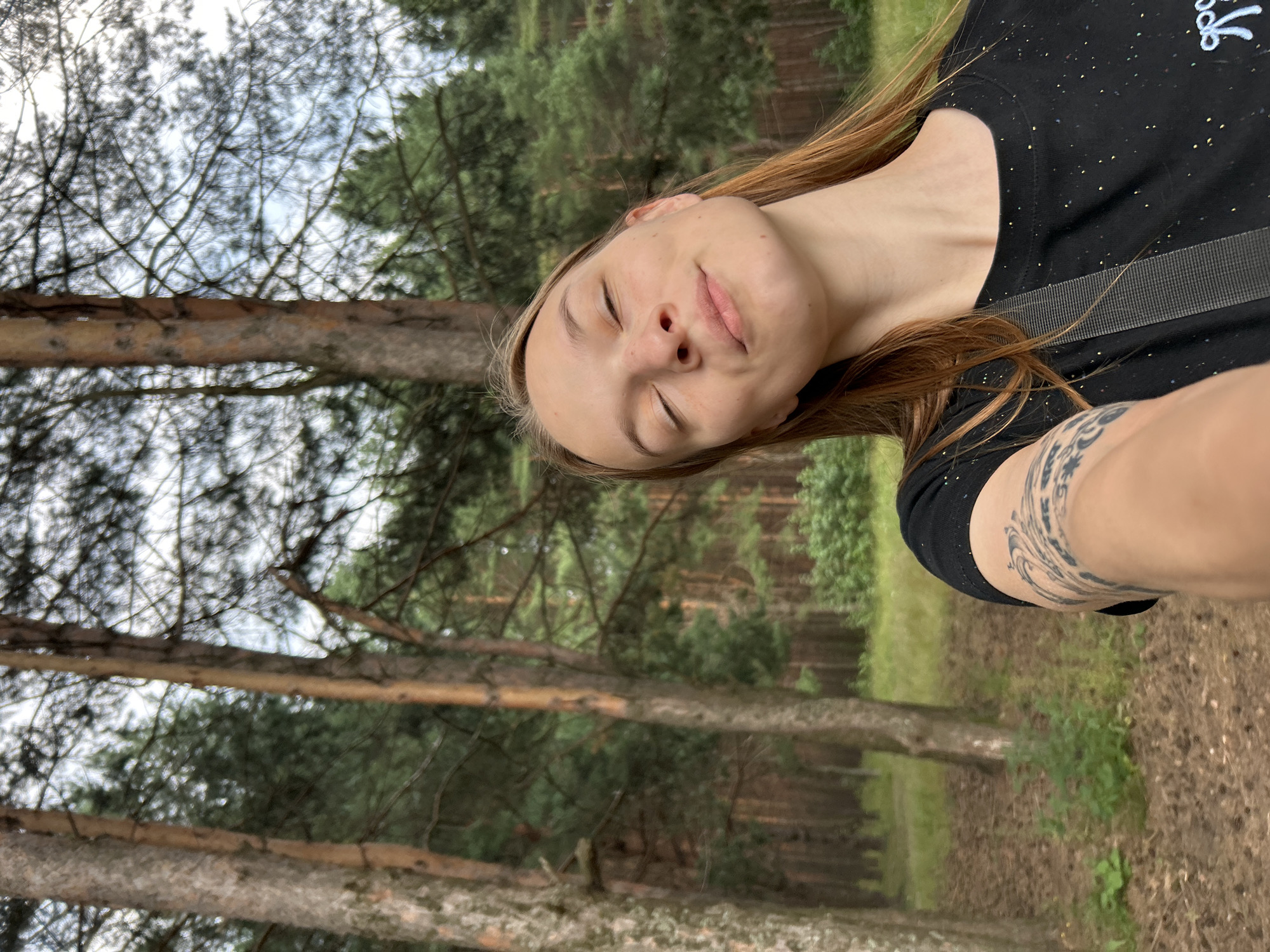 In a forest setting, a young woman with blonde hair and closed eyes smiles with her head tilted back. She is wearing a black t-shirt with a strap across her chest and has a tattoo on her left arm. The background consists of tall pine trees, green foliage, and a cloudy sky. The location is the city of Chaikovsky, Russia.