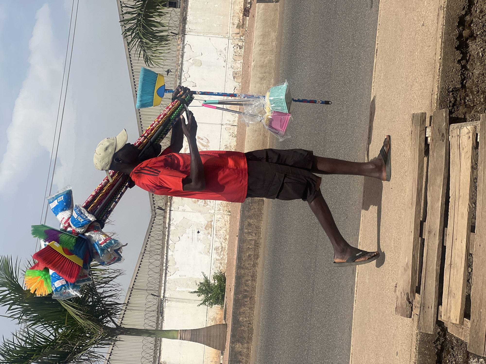 A man is walking along the side of a road in Barakin Lerawi, Nigeria, carrying a large bundle of brooms and mops over his shoulder. He is wearing a red t-shirt, black shorts, a light-colored cap, and sandals. He appears to be looking at or adjusting the brooms on his shoulder. The road is paved, and a sidewalk runs alongside it with wooden planks laid in a row. A white wall with a metal fence along the top runs parallel to the road. Palm trees and other vegetation are visible in the background. The sky is partly cloudy.