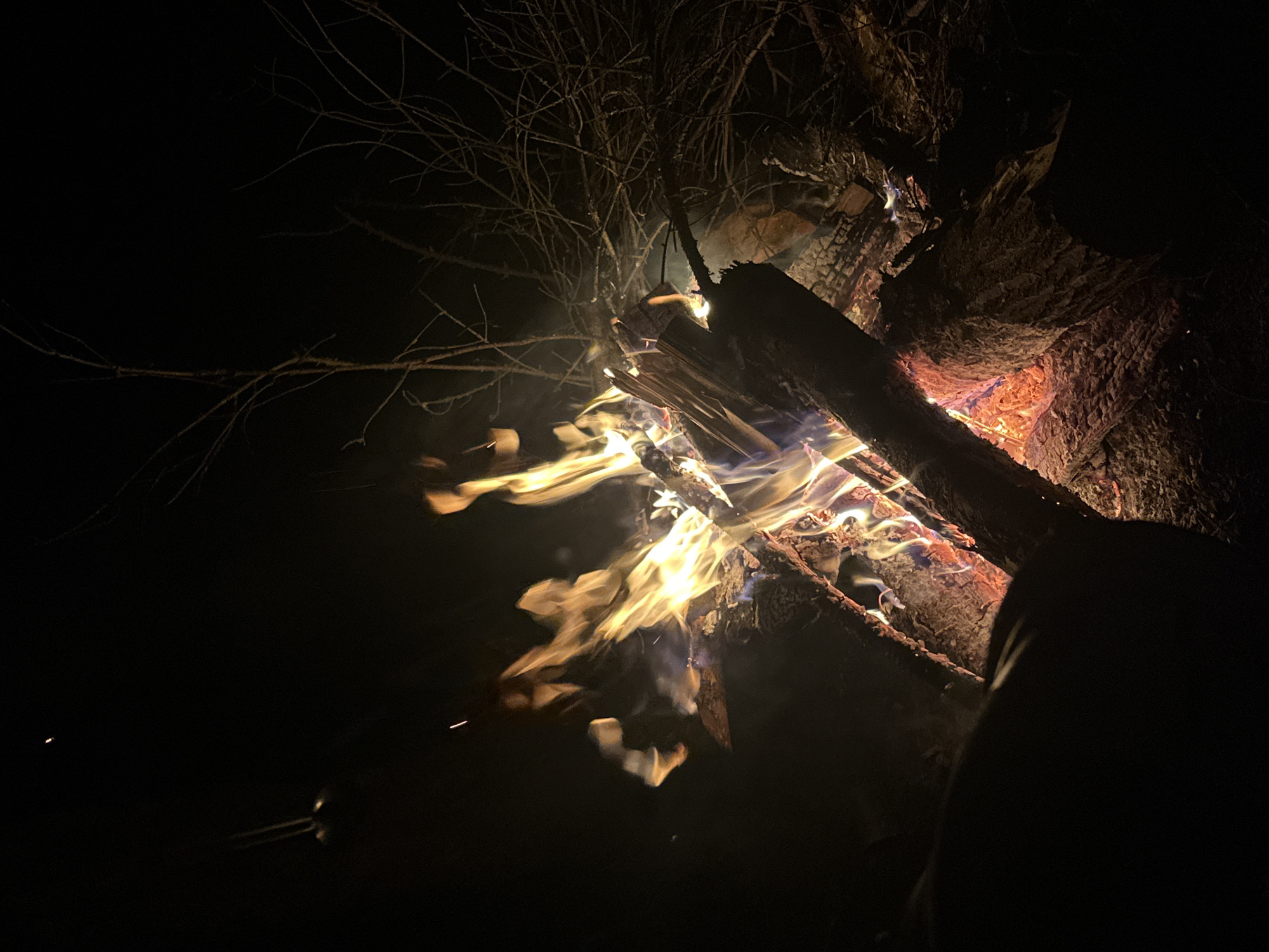 At night in Папярнянскі сельскі Савет, Belarus, a campfire burns, consisting of several logs arranged to support the flames. Thin branches rise above the fire. Part of a person's body is visible in the lower right.