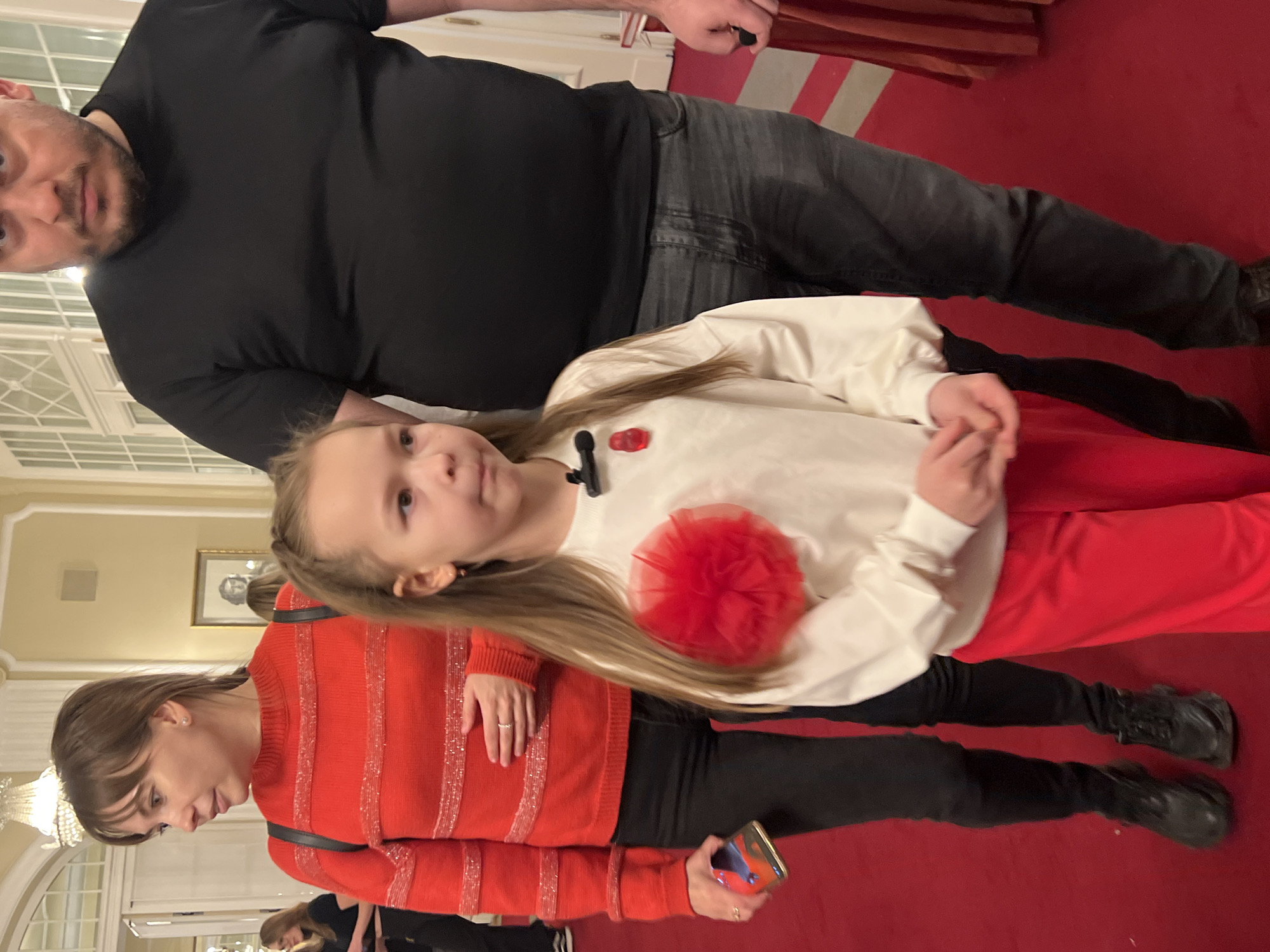 Three individuals are in the frame, in what appears to be an ornate indoor setting. A man in a black shirt and gray pants is positioned near the top of the frame. A woman is present and wearing a red-striped shirt and black pants. A young girl with light hair, wearing a white shirt, a decorative red flower, and red pants, is in the middle. All three are on a red carpet. In the background, there's a glimpse of a building's interior with windows and architectural details, which suggest that the location might be the Yusupov Palace on the Moika River in Saint Petersburg, Russia.
