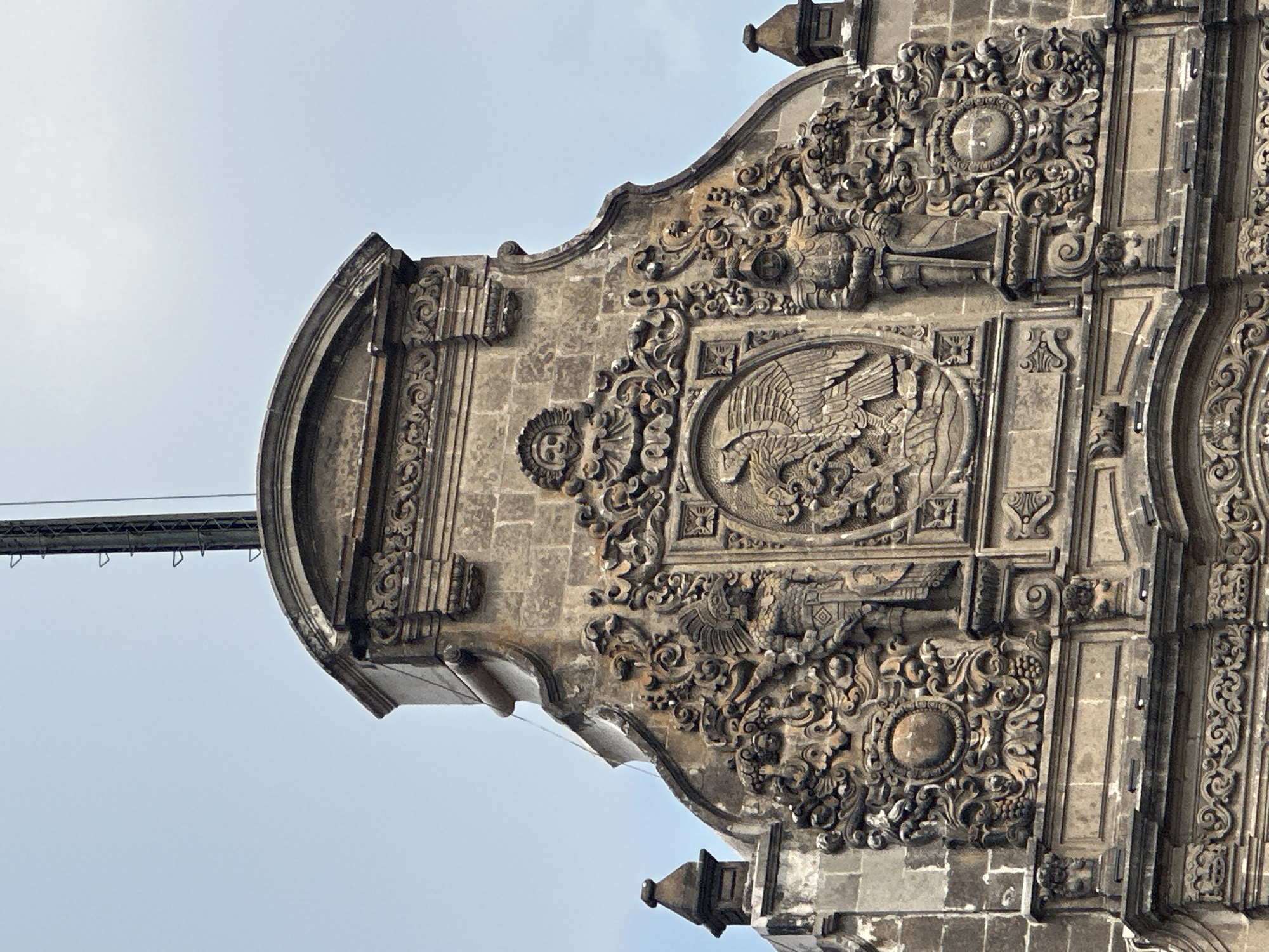 The image shows the upper facade of a building in Mexico City, Mexico. The facade is ornately decorated with carvings. Prominently featured is a circular relief depicting an eagle, possibly the Mexican coat of arms. Other carvings feature floral and geometric patterns. The facade is made of light-colored stone. A small portion of a cable is visible in the background against a pale blue sky.