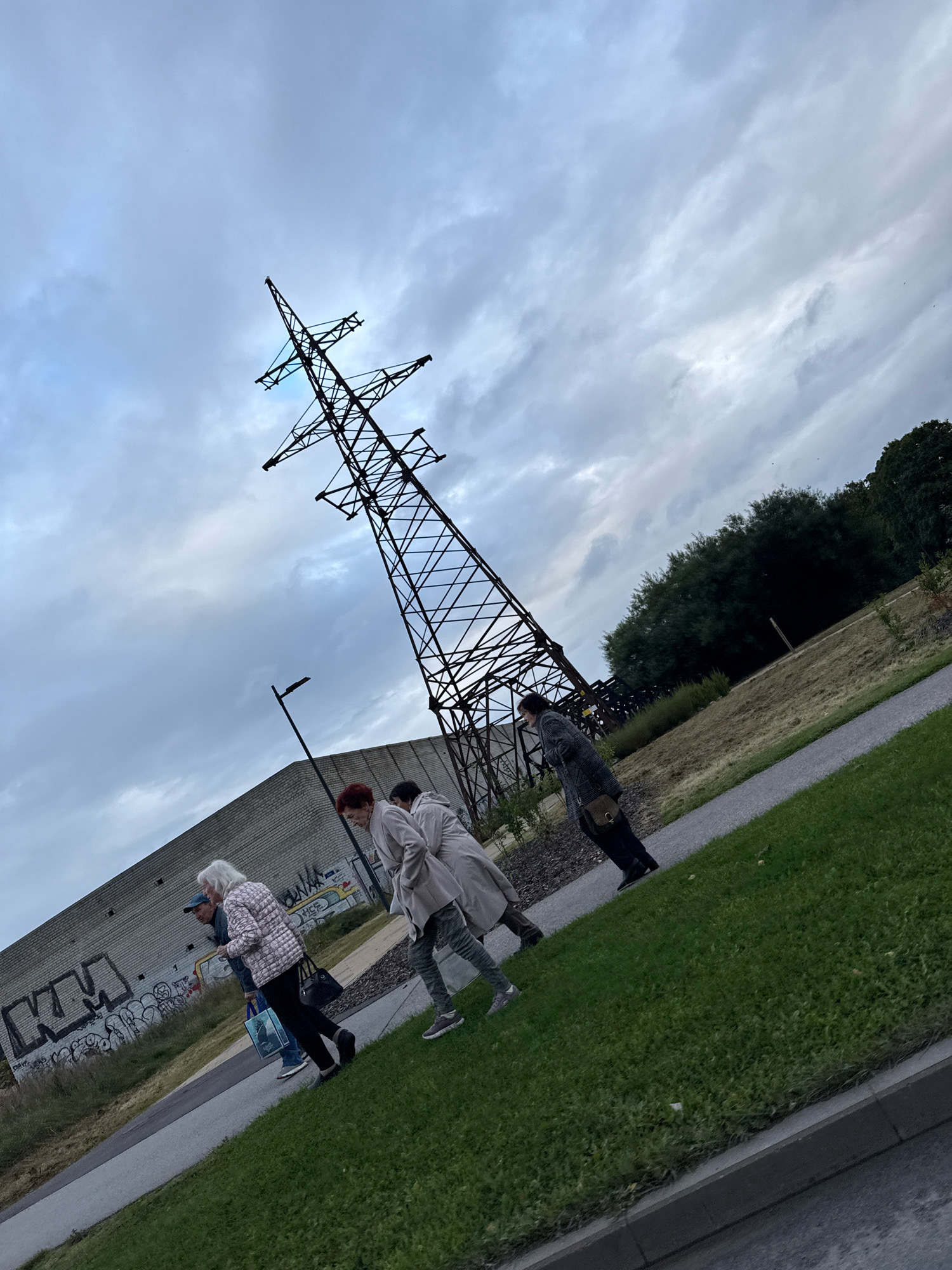 The image depicts an outdoor scene with a tilted perspective, suggesting the camera is angled upwards or the ground is uneven. Dominating the frame is a tall, dark metal electricity pylon, its lattice structure stark against a cloudy, overcast sky. To the left of the pylon, a long, grey brick building with graffiti on its lower section is visible. A single black lamppost stands between the building and the pylon.
In the foreground and mid-ground, four individuals are walking along a paved path that runs parallel to a strip of green grass. The first person from the left, an elderly woman with white hair, is wearing a light-colored jacket with a patterned design and black pants. She carries a dark handbag. Next to her is a person wearing a blue cap and a darker jacket. A woman with short red hair, wearing a light-colored coat and grey pants, is walking in front of them. The last person visible is wearing a dark coat and black pants, carrying a brown shoulder bag. They appear to be walking away from the viewer, towards the right side of the image.
The environment suggests an urban or semi-urban setting, possibly an industrial area transitioning into a park or recreational space, given the presence of the pylon, industrial building, and the path with grass and trees in the background. The overall mood is somber due to the cloudy sky and the muted colors. The location is identified as being in the city of Tallinn, Estonia.