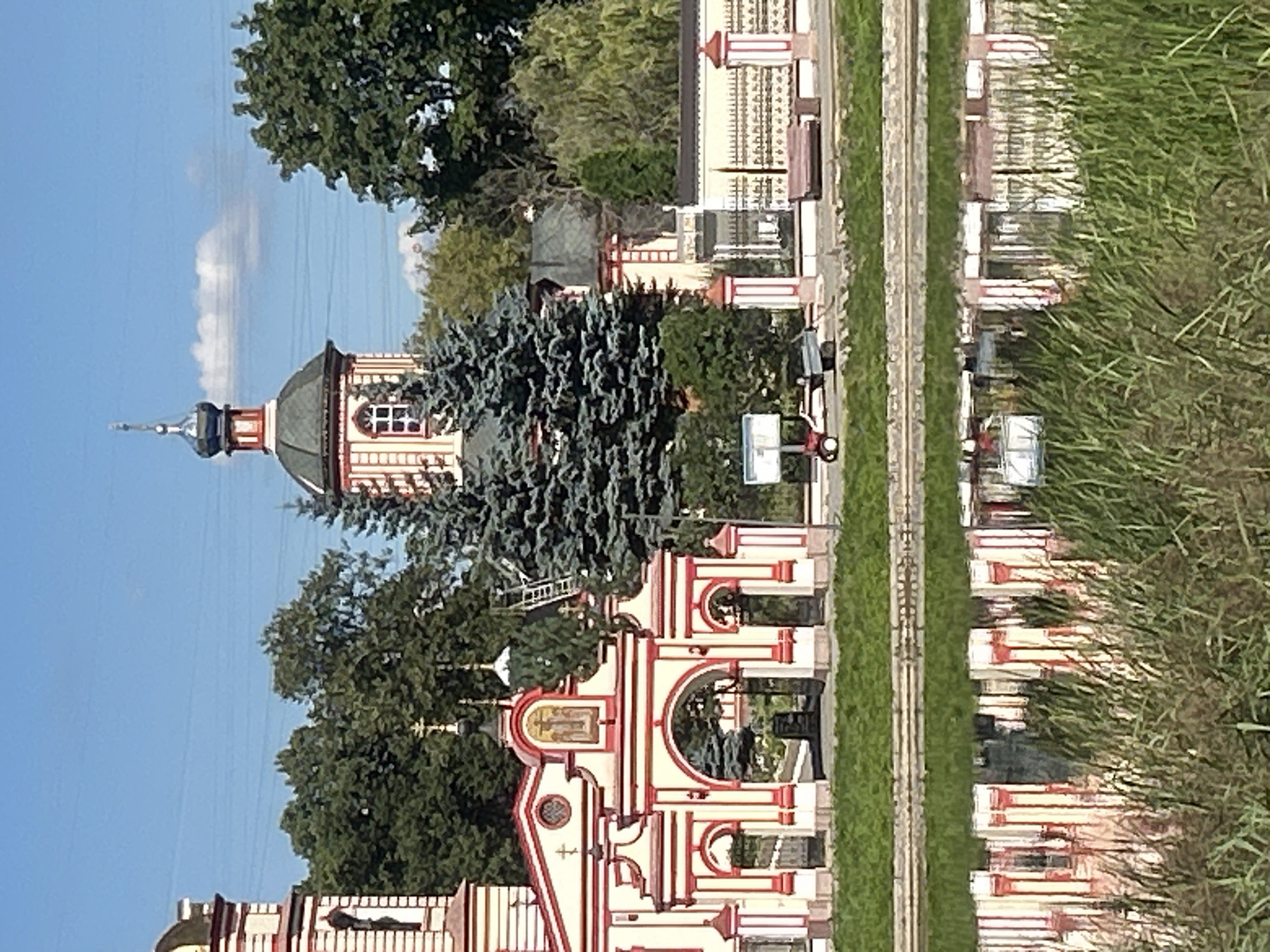This image depicts the facade of a colorful Orthodox church, likely a monastery, in Moscow, Russia. The architecture is ornate, with white walls accented by red trim and gilded domes. A prominent feature is the arched gateway with decorative columns leading into the complex. In the background, lush green trees and a tower with a spire rise against a clear blue sky. A body of water in the foreground reflects the church's architecture and the surrounding greenery. The scene suggests a tranquil, possibly historical, religious site.