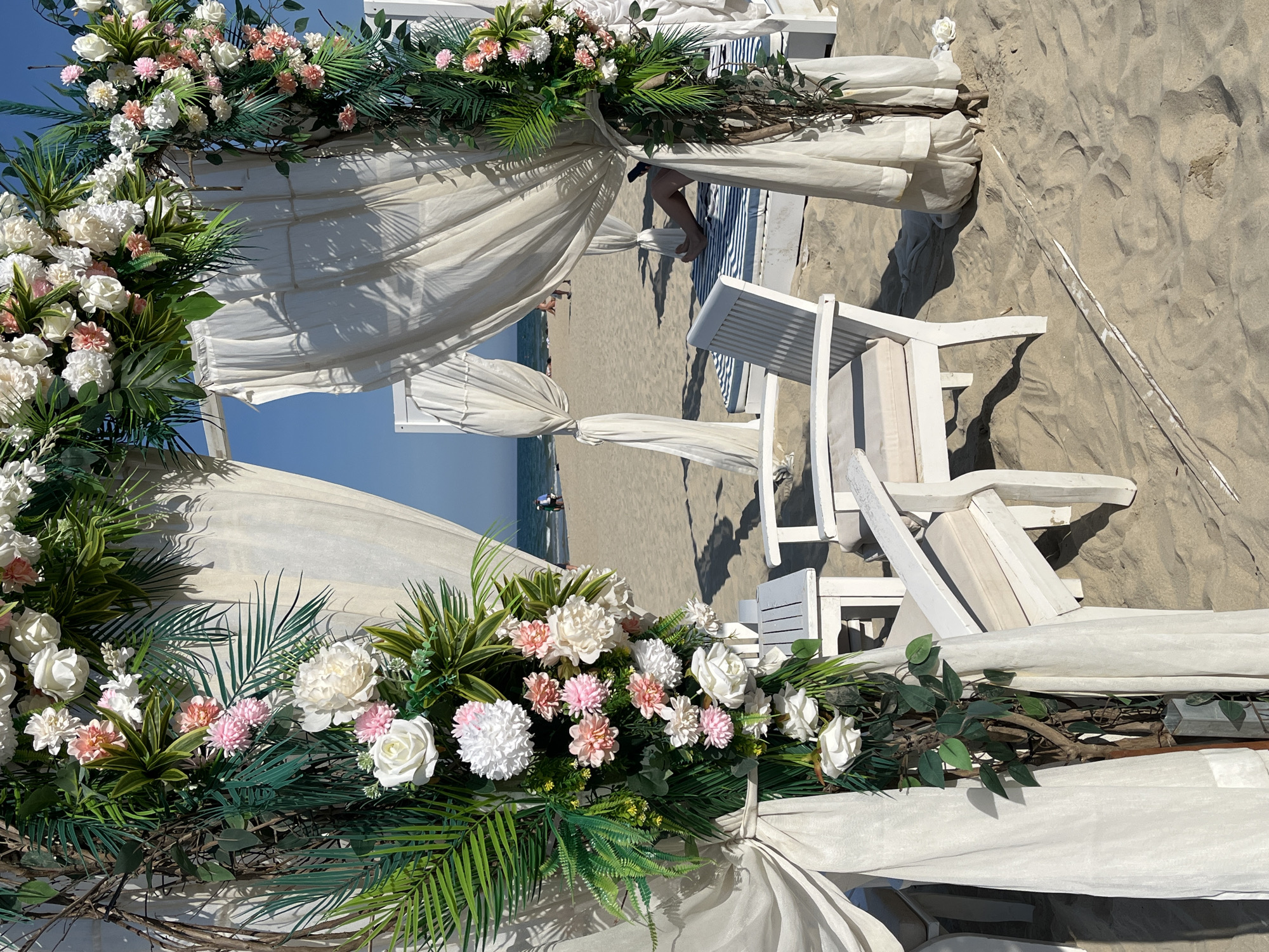 The image depicts a beach scene in Đà Nẵng, Vietnam. A white wooden structure, possibly a wedding arch or decorative canopy, is prominently featured. It is adorned with arrangements of white and pink flowers and green foliage. White drapes hang from the structure. The backdrop includes a sandy beach, the ocean, and a clear sky. There are a few figures visible in the distance along the shoreline. White wooden beach chairs are also present.