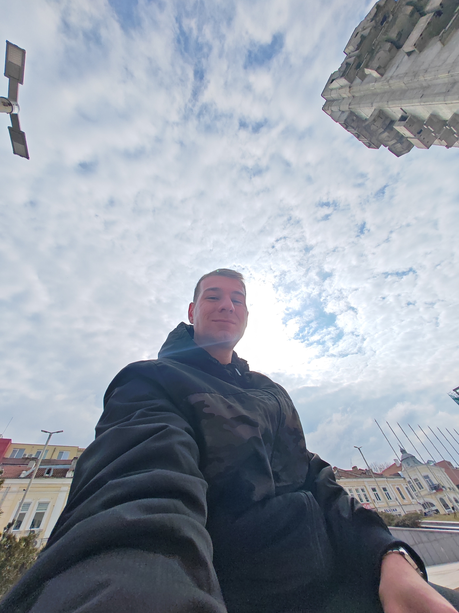 A low-angle, wide-field-of-view image captures a young adult male taking a selfie outdoors in Shumen, Bulgaria. The subject, positioned centrally, is smiling and looking directly at the camera. He wears a dark, hooded jacket, featuring a subtle camouflage pattern across the shoulders and upper arms. A metallic watch or bracelet is visible on his right wrist.
The setting is an urban environment under a partly cloudy sky, with patches of bright blue and a visible bright sun glare. In the upper left corner, a modern street lamp with two rectangular LED luminaires is visible. To the upper right, a significant portion of the Monument to 1300 Years of Bulgaria, a large, angular Brutalist concrete structure, is prominent.
Behind the male, to the left, are facades of multi-story buildings, including a light-colored structure with numerous windows, partially obscured by bare tree branches. A red building is visible beyond it. To the right, another ornate, light-colored historic building featuring a distinctive dome-like roof section and multiple windows is present. A partial sign or banner on this building displays the Cyrillic text "ОБЩИНА". Further right, a series of thin, vertical poles, possibly flagpoles, are visible. The subject appears to be seated or positioned low on a concrete platform or wall, with an additional low concrete barrier visible in the lower right portion of the frame.