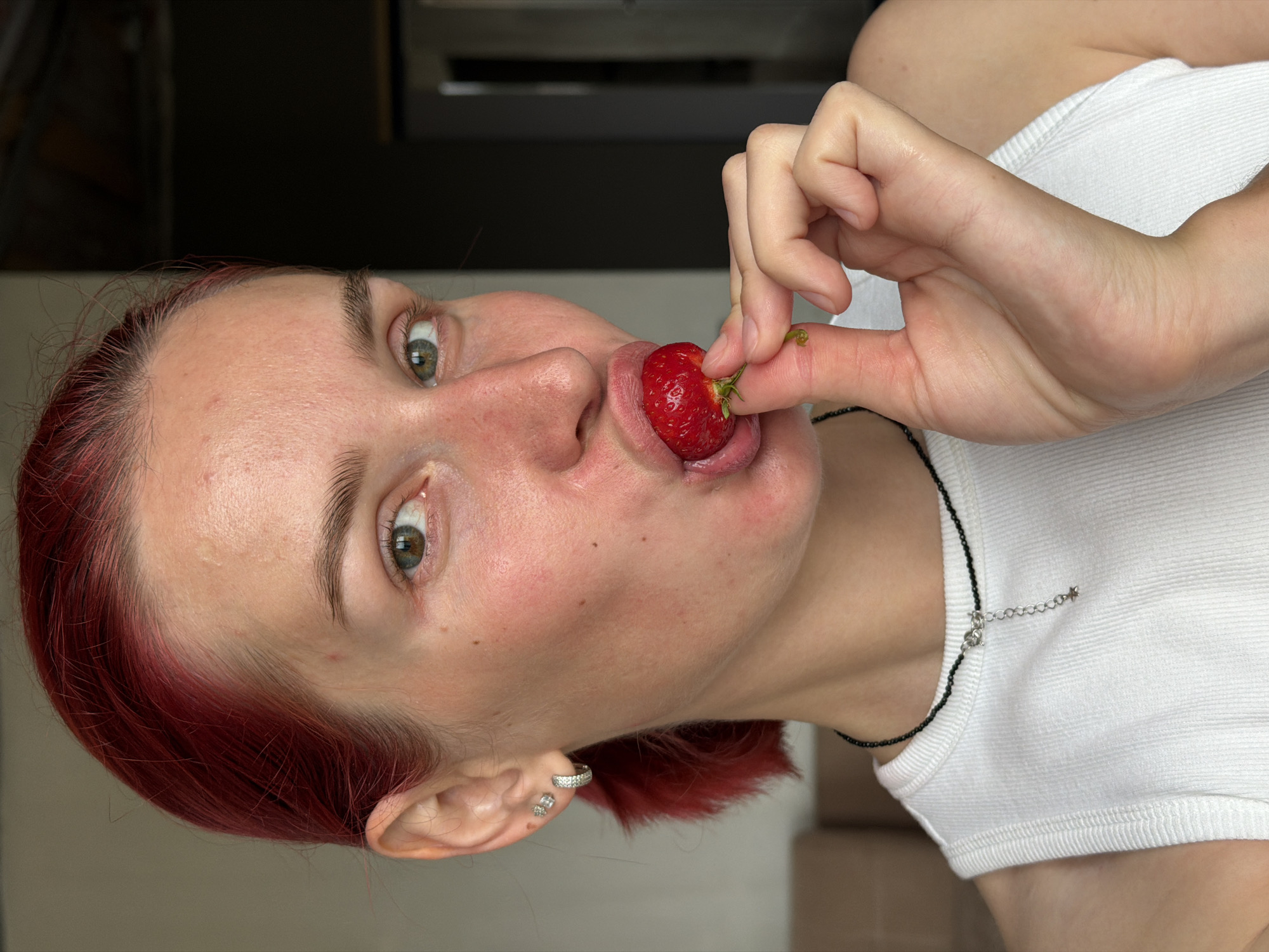 A woman with bright red hair and light green eyes is captured in a close-up shot, holding a strawberry to her lips. She is wearing a white tank top and a black beaded necklace with a small silver charm. The woman has several piercings in her left ear. The background is blurred, showing a glimpse of a room with muted tones and furniture. There is no identifiable location information about Chaikovsky, Russia in the image.