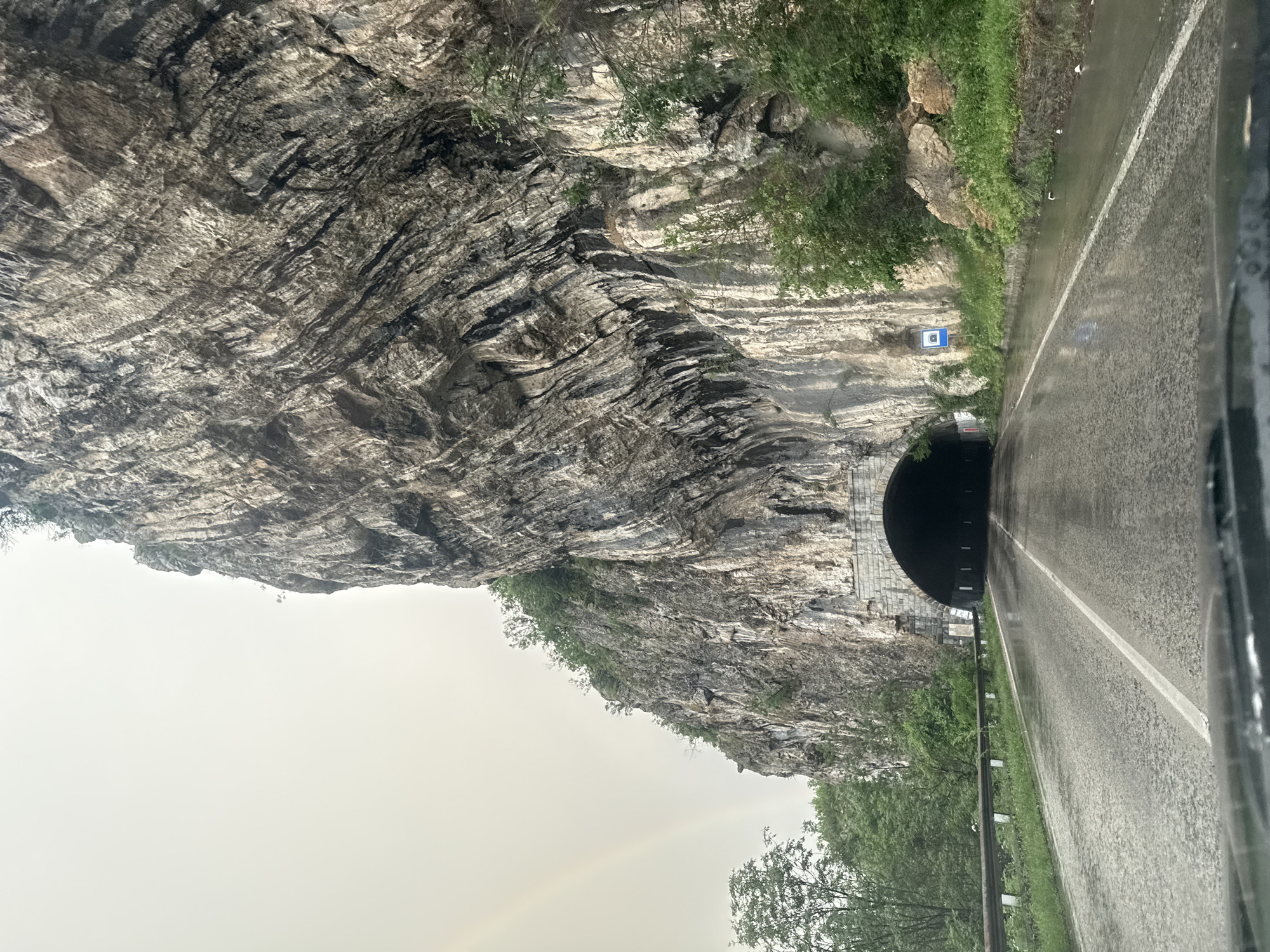 A road passes through a tunnel in the rock face near Gesha, Bulgaria. The road is wet, with a white line marking the edge. A rock face looms over the road and tunnel, while green vegetation is visible on the edge of the rock face and along the road. A pale rainbow appears in the lower left corner of the image.