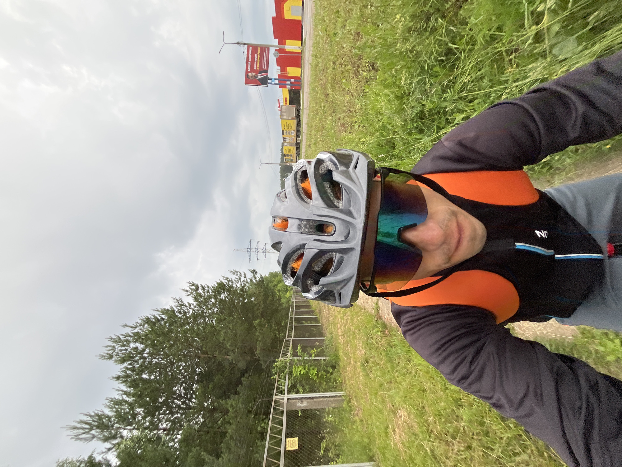 The image shows a first-person perspective of a cyclist wearing a black helmet and blue-tinted sports glasses, with an orange backpack visible. The cyclist is on a dirt path, with tall green grass and trees lining the left side of the path, bordered by a chain-link fence. In the background, there are power lines, a billboard featuring a man in a suit, a row of yellow and red buildings, and several yellow semi-trailer trucks. The sky is overcast and cloudy. The scene appears to be on the outskirts of the city of Chaikovsky, Russia, possibly near an industrial or commercial area given the buildings and trucks.