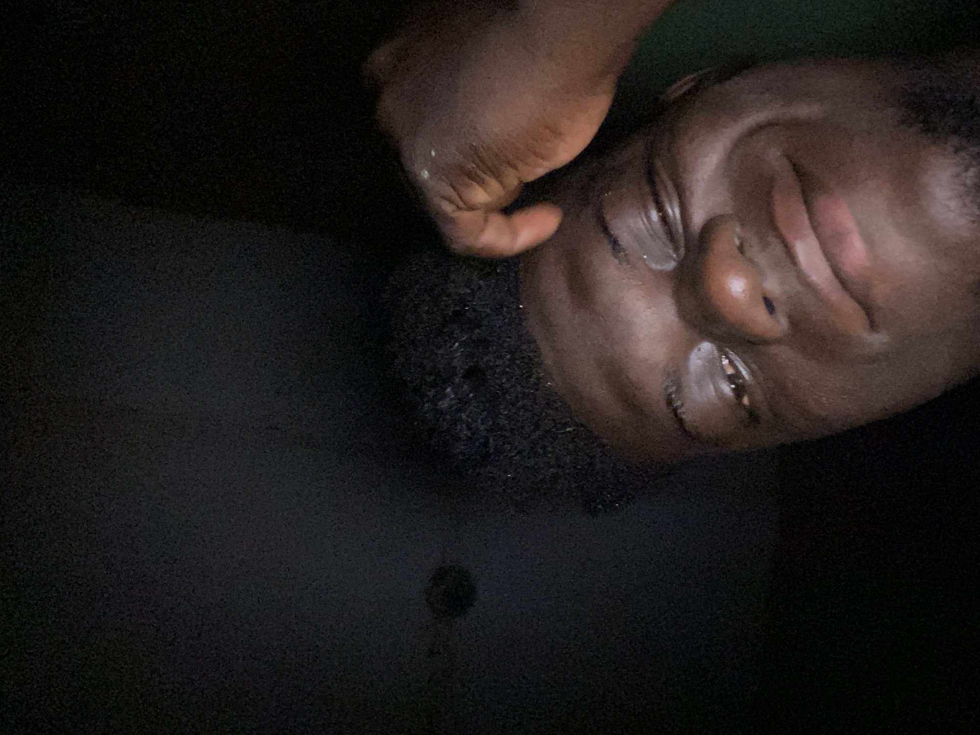 A dark-skinned man is positioned with his head tilted to the right. His hand is near his face, with some white substance possibly on his fingers. The background is dark, with minimal detail visible, except for a dark spot or object. The location is unspecified, with the city listed as Unknown, Unknown.