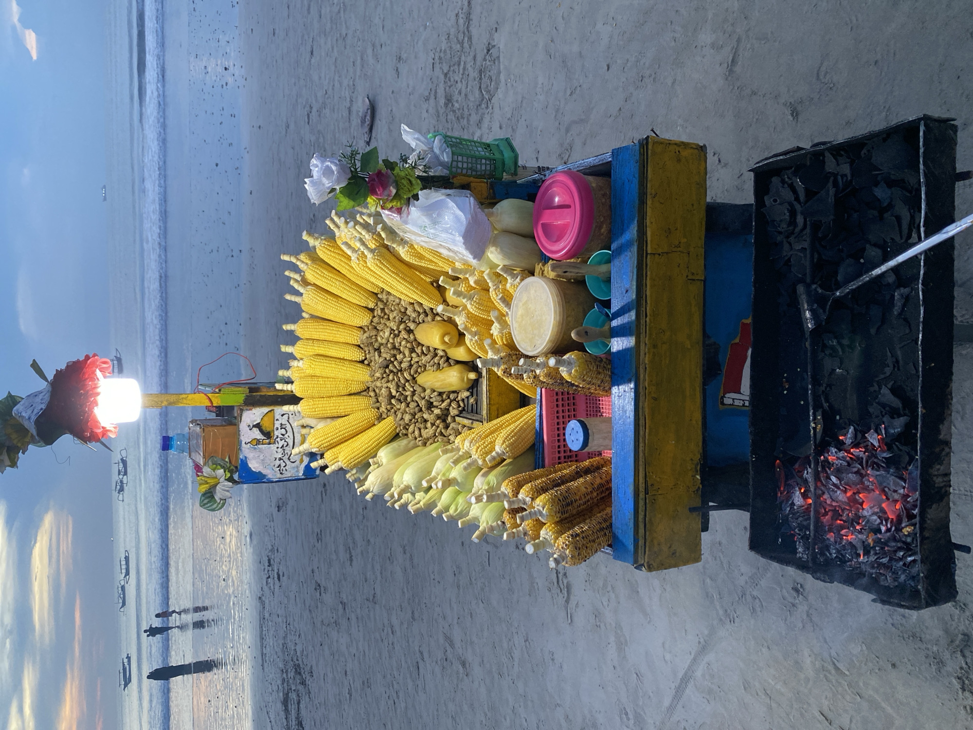 The image captures a corn and peanut vendor's cart on a beach in Kuta Selatan, Indonesia. The cart is filled with rows of corn on the cob, both raw and grilled, and piles of peanuts. Various containers and food preparation items are also present. A charcoal grill with glowing embers is attached to the cart, suggesting the corn is grilled on-site. In the background, the beach is visible, with calm waves meeting the shore. Distant figures of people are silhouetted against the sky, and boats are present near the horizon. The lighting indicates either sunrise or sunset.