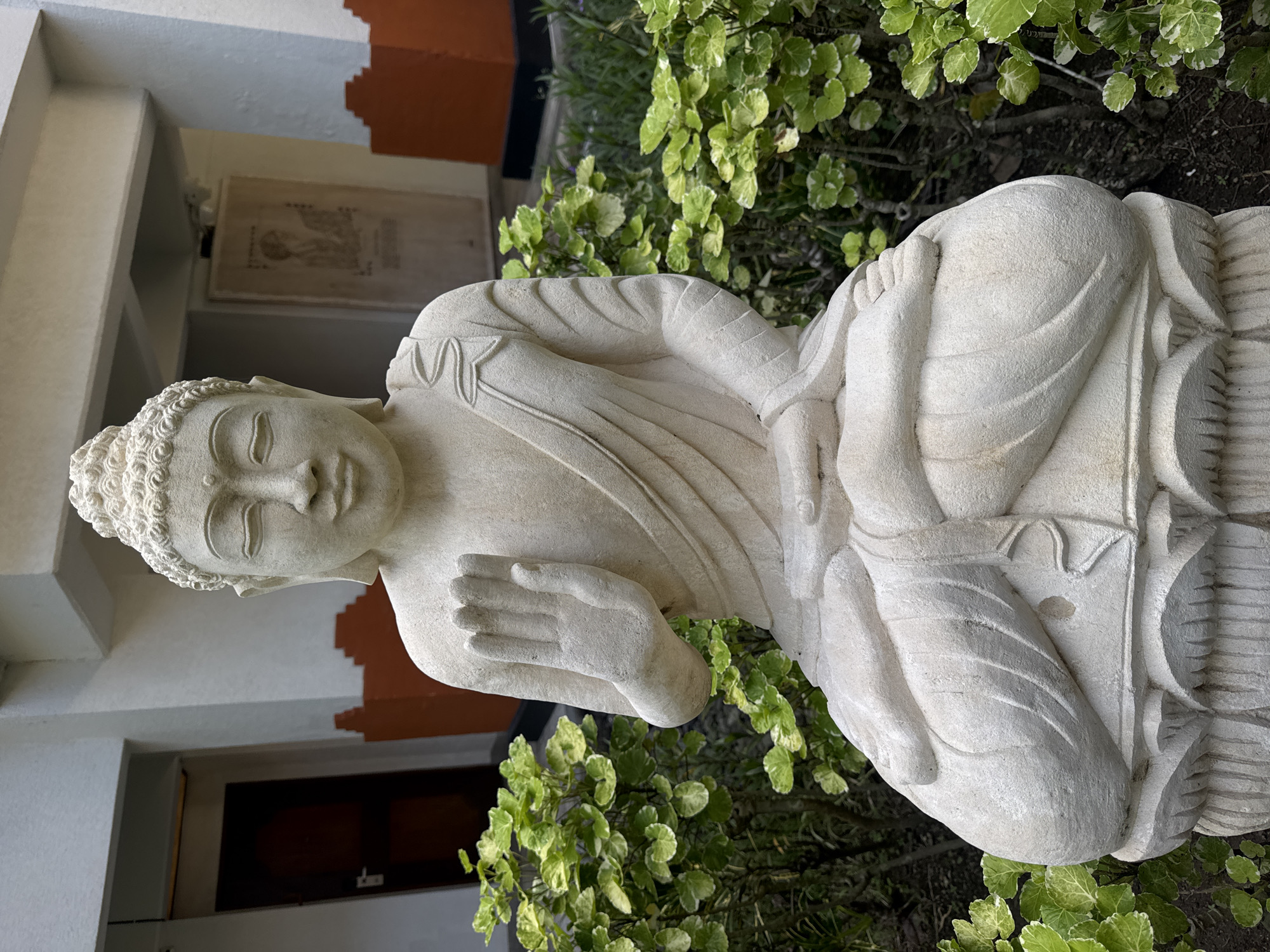 A detailed description of the image follows:
**Subject:** The image features a stone statue of Buddha, placed outdoors amidst vegetation.
**Setting:** The statue is positioned within a garden or courtyard setting. The surroundings include green plants with variegated leaves. Architectural elements are visible in the background, suggesting an urban setting.
**Location:** Although the image does not offer specific details, the prompt indicates a location in Kuta Selatan, Indonesia.
**Objects:** The primary object is the stone Buddha statue, exhibiting intricate carvings and details. Other objects include plants, and architectural structures in the background.
**Environment:** The environment appears to be outdoors. The presence of plants indicates a garden or landscaped area.
**People:** There are no people visible in the image.
**Actions/Interactions:** The scene depicts a static, serene arrangement. The statue serves as a focal point, with the surrounding vegetation providing a natural backdrop. The image captures a moment of quiet contemplation or reverence.